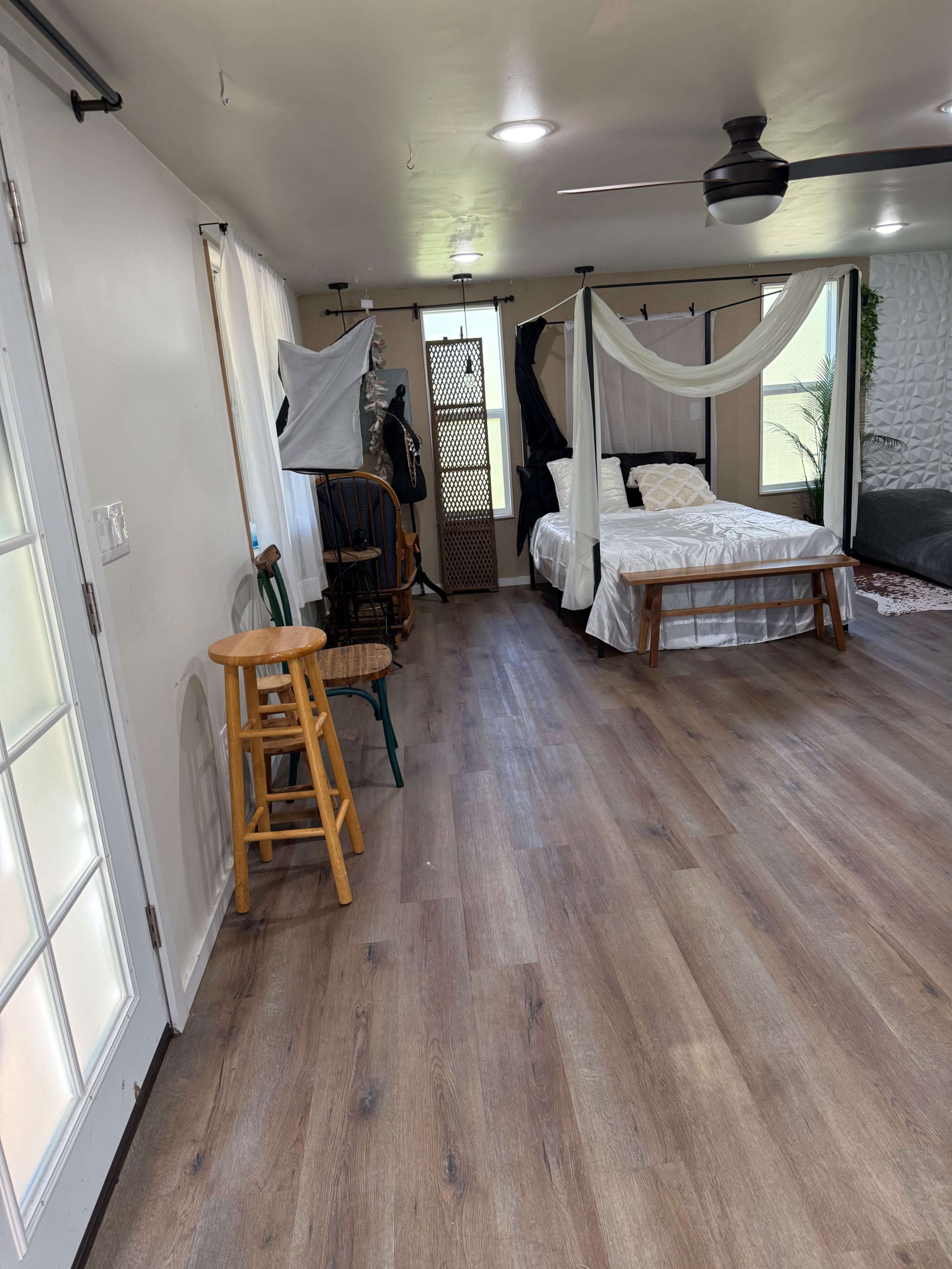 The image shows a spacious, well-lit room featuring a bed with draped fabric, a stool, and various photography equipment arranged along the walls.