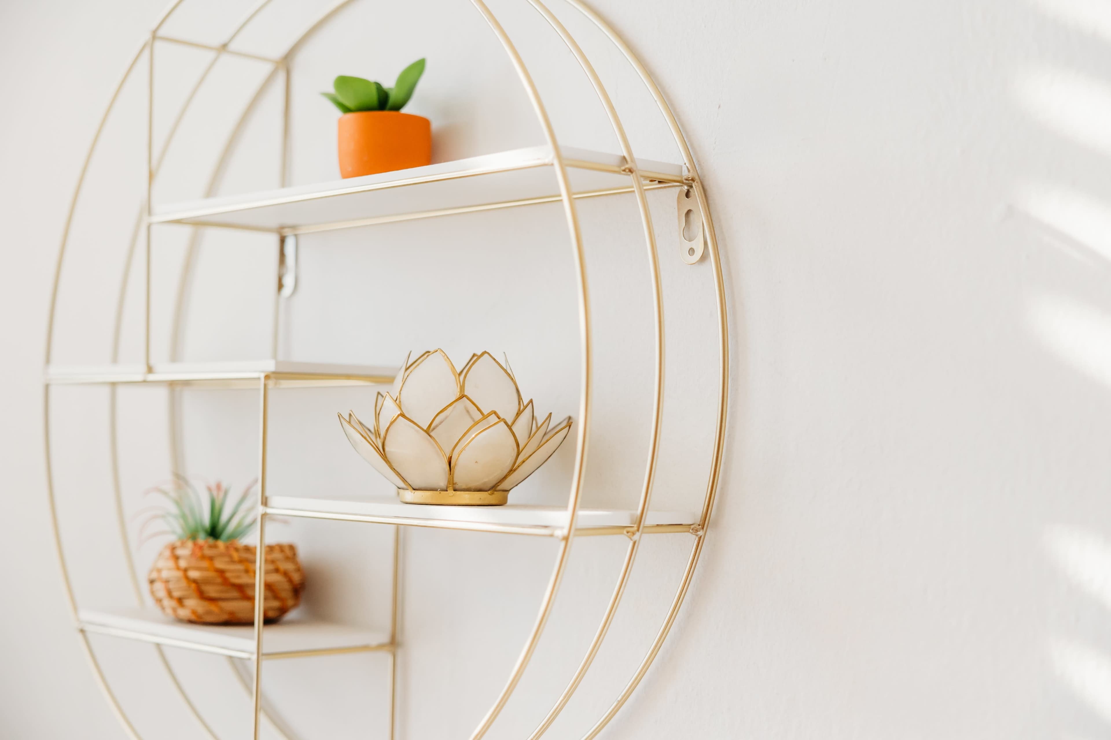 A circular gold shelving unit mounted on a white wall displays a small potted plant, a woven basket, and a decorative lotus flower.