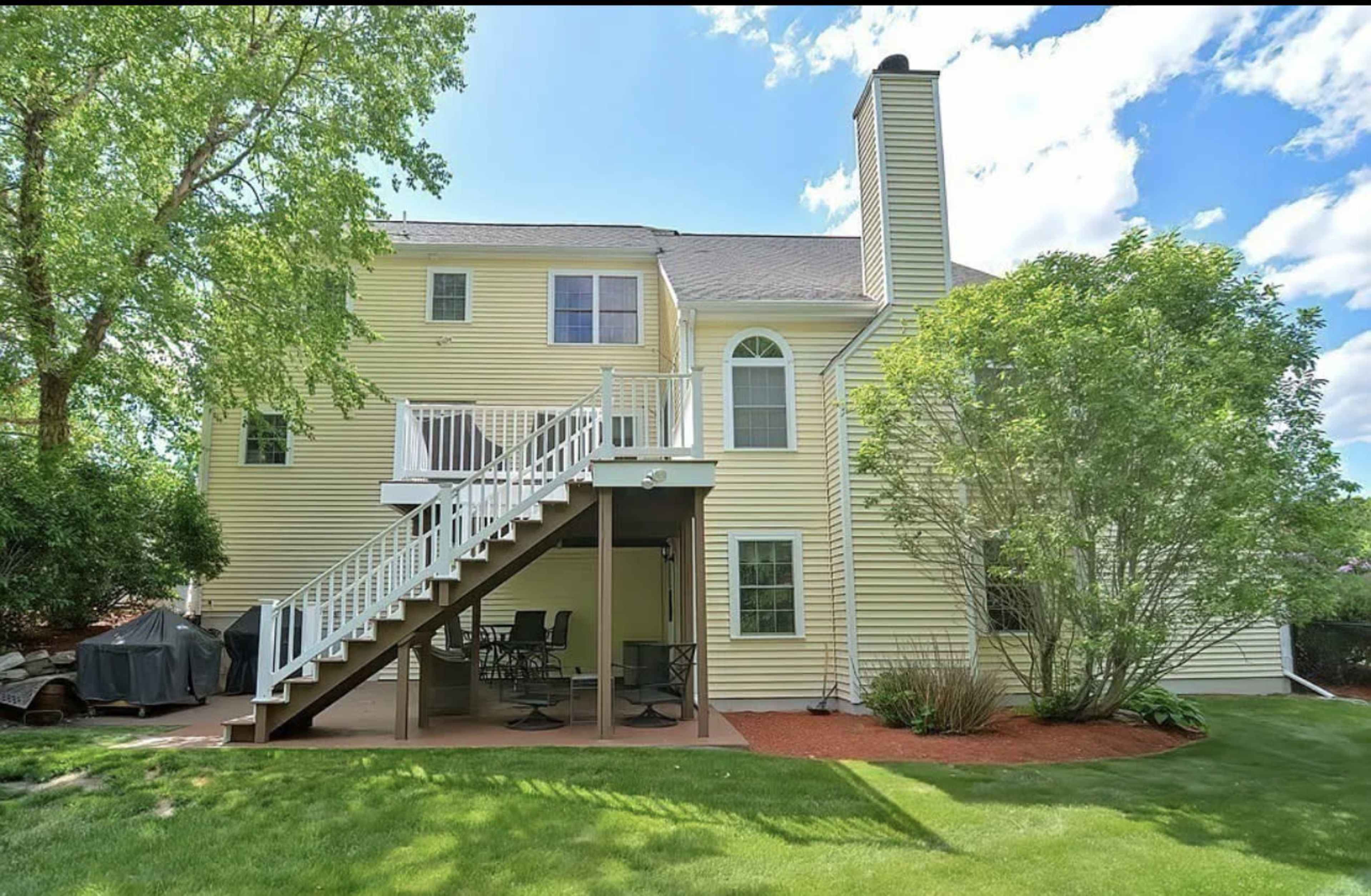 The back exterior of a yellow two-story house with a deck and patio area, surrounded by lush greenery.