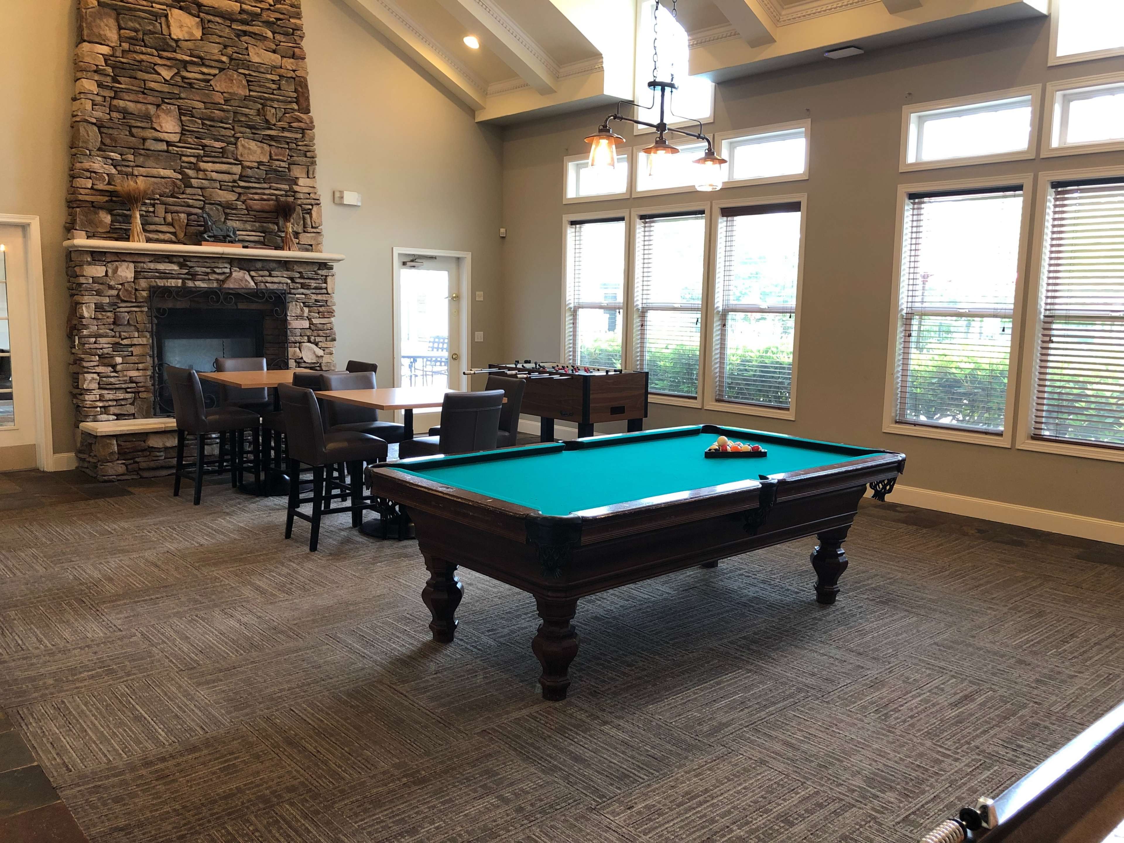 The image shows a recreational room featuring a green pool table, a stone fireplace, and several windows allowing natural light.