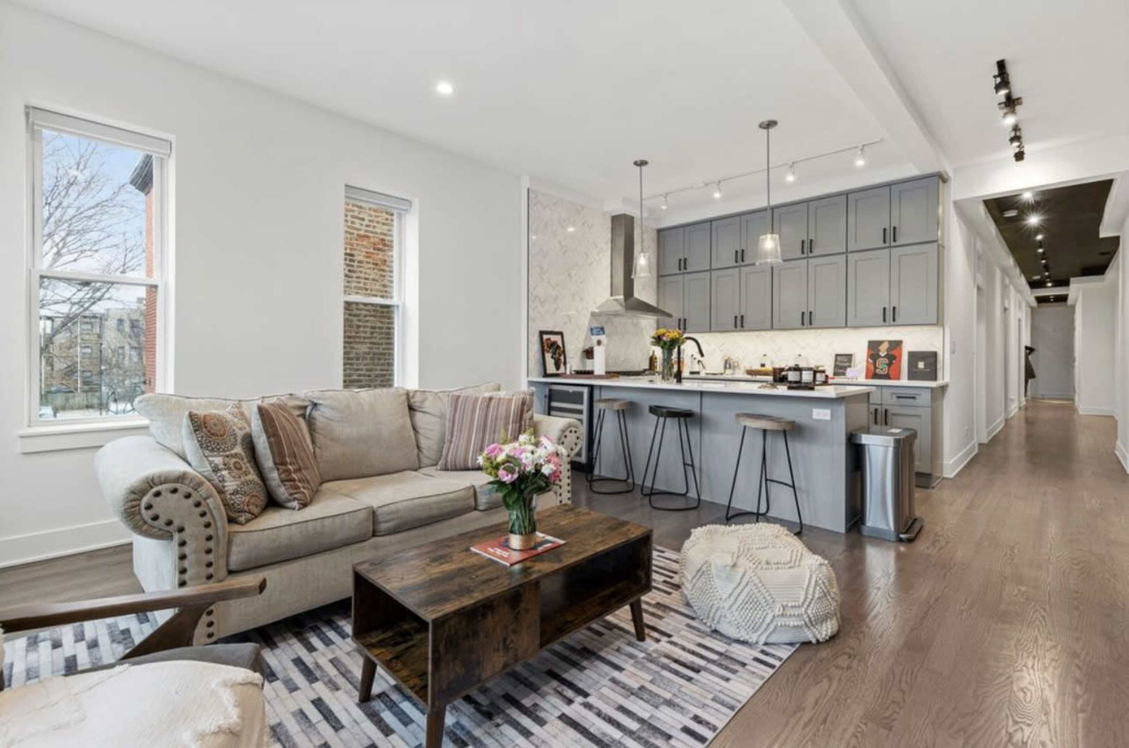 The image shows a modern living area featuring a beige sofa, a wooden coffee table, and a kitchen with gray cabinetry and a marbled backsplash.