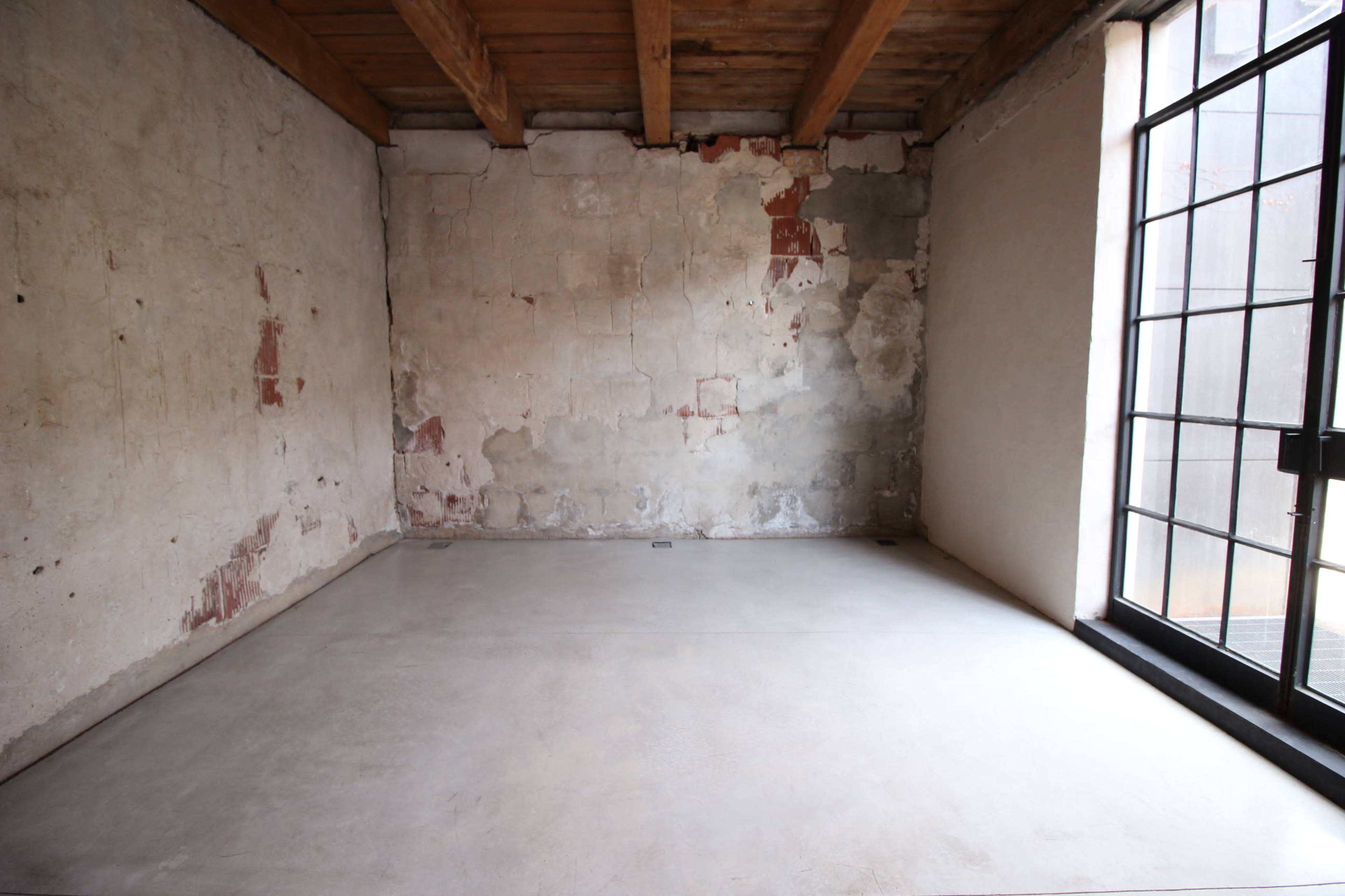 An interior room with bare walls, a concrete floor, and large windows.