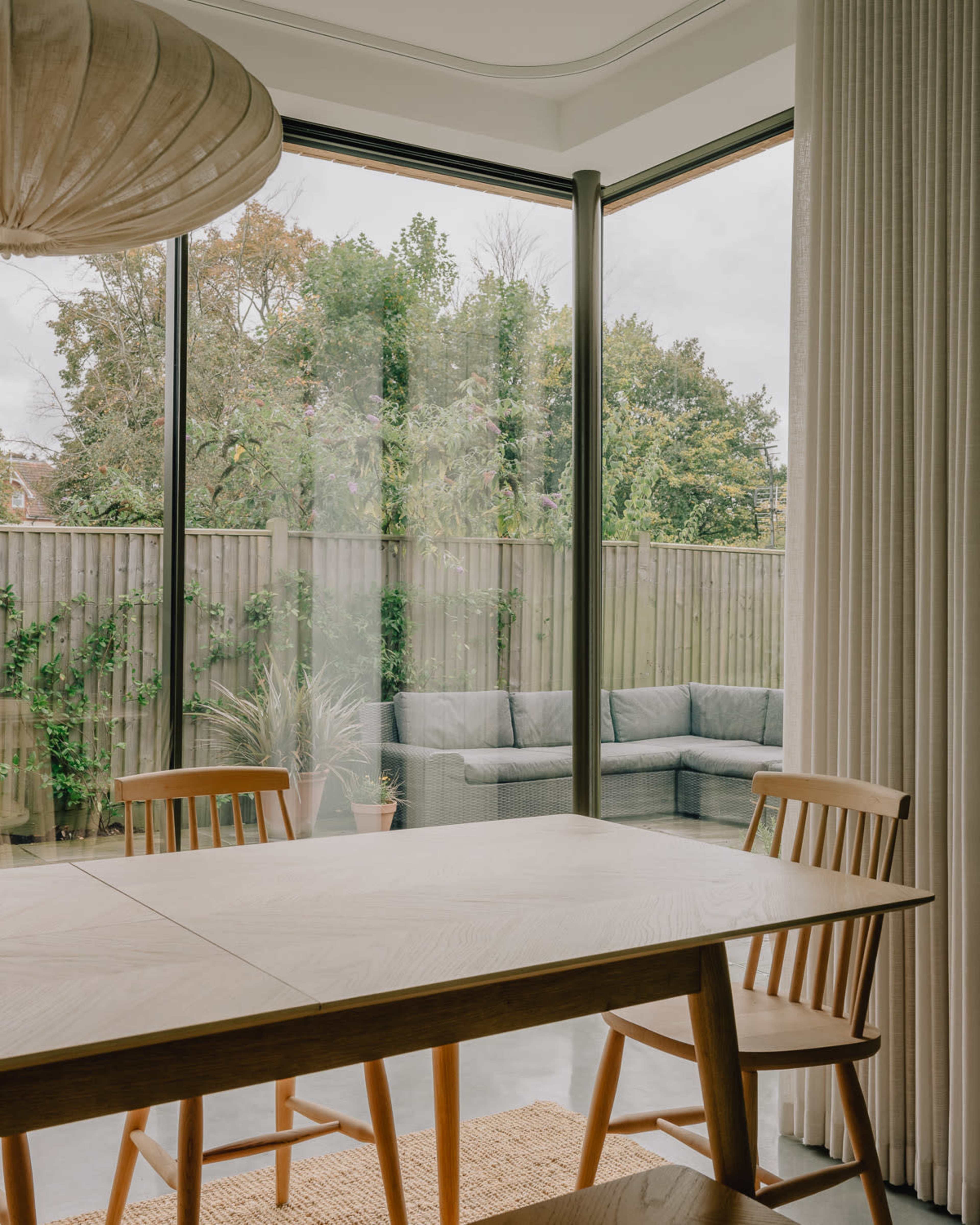 A dining area features a wooden table and chairs, with large windows offering a view of a landscaped outdoor space with a seating area.