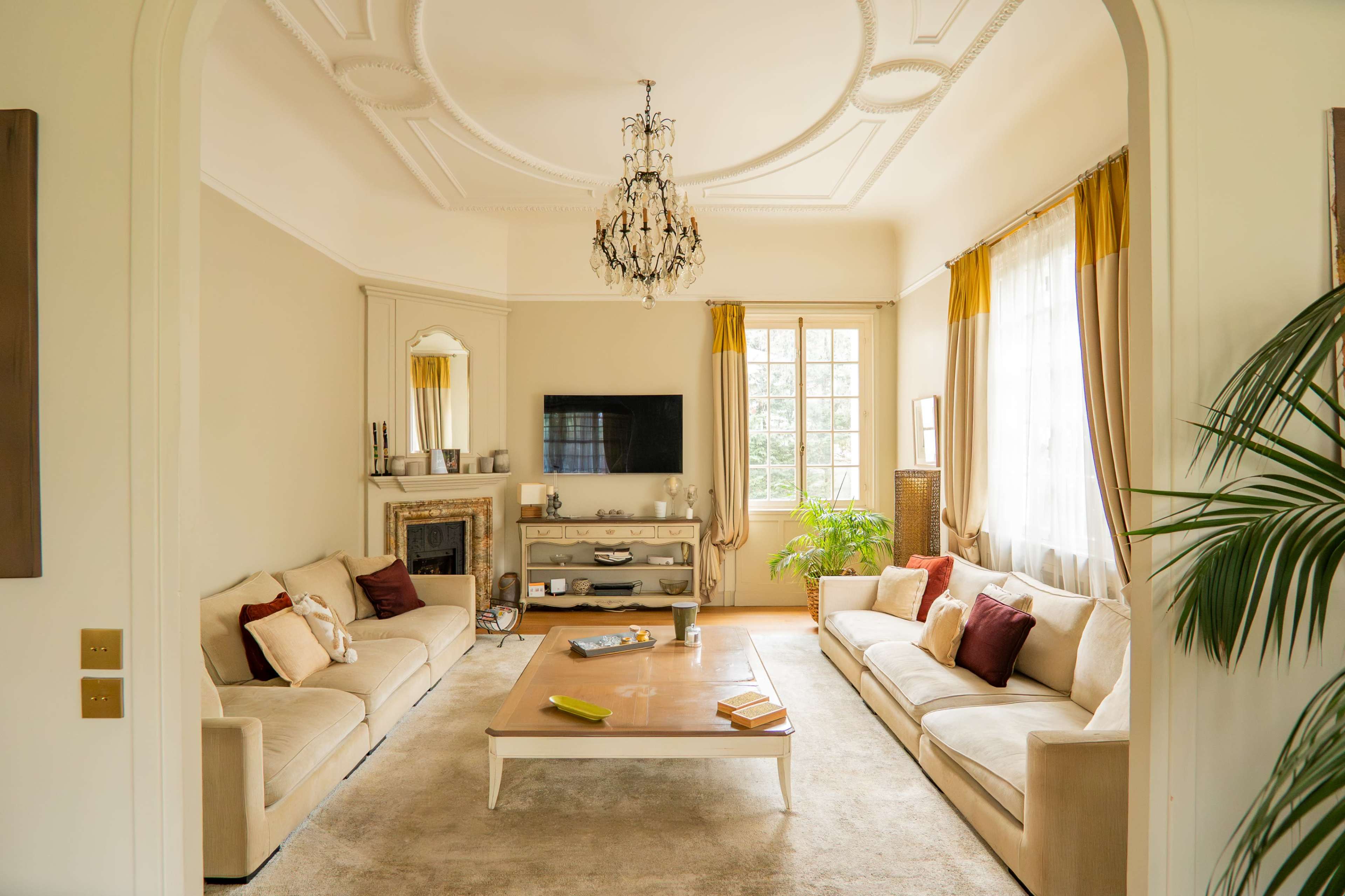 The scene features a well-lit living room with two sofas, a coffee table in the center, a television on the wall, and large windows allowing natural light to enter.