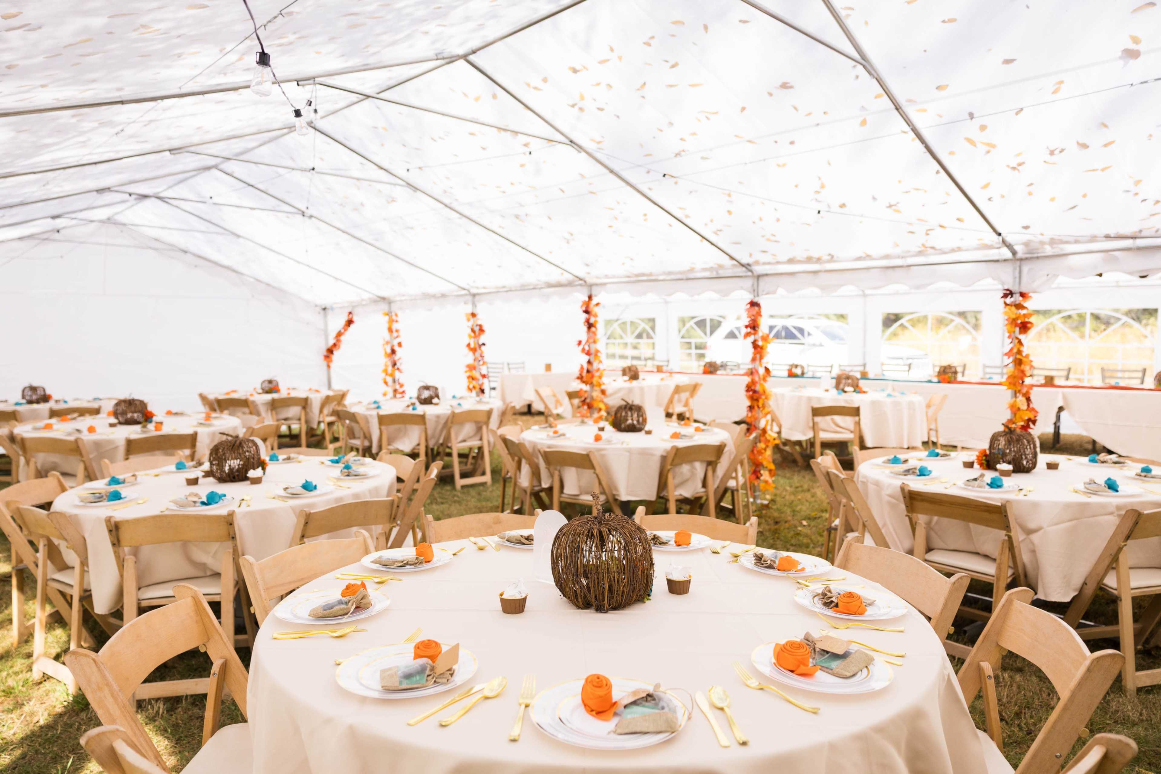 A large tent is set up for an event, featuring multiple round tables adorned with white tablecloths, orange pumpkins, and colorful decorations.