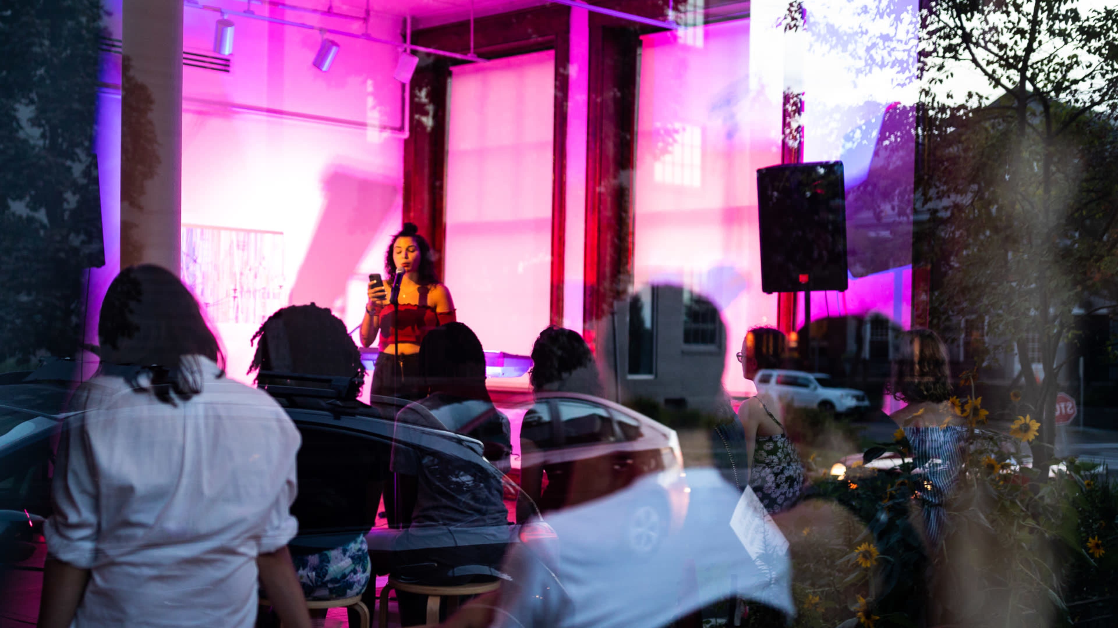 A performer stands on stage, speaking to an audience seated in front of her, with colorful lighting illuminating the space.