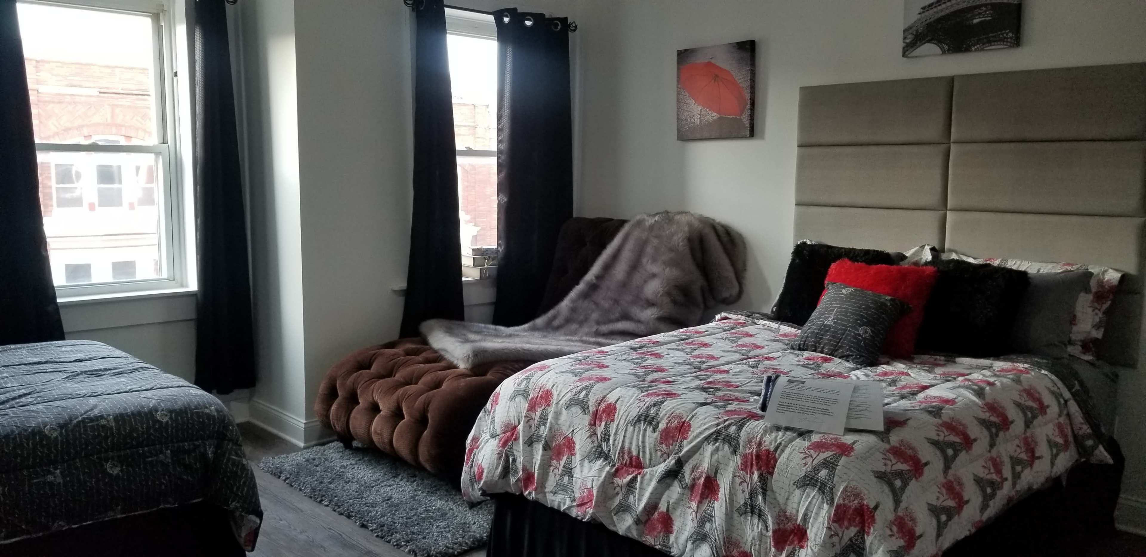 A bedroom with two beds, one with a patterned comforter and the other with a darker, solid cover, along with large windows and dark curtains.