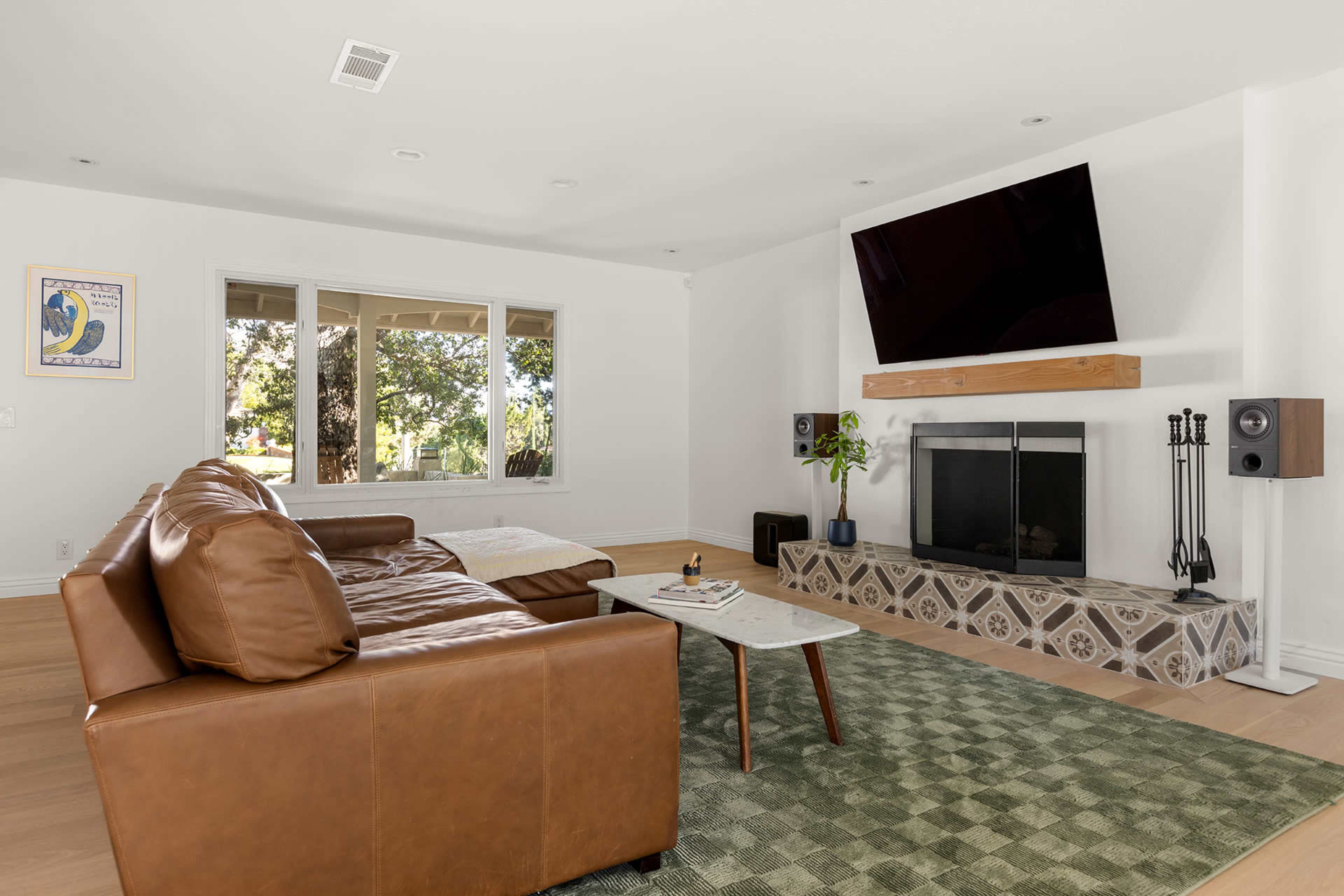 The living room features a brown leather sofa, a wall-mounted TV above a fireplace, and a patterned tile surround.