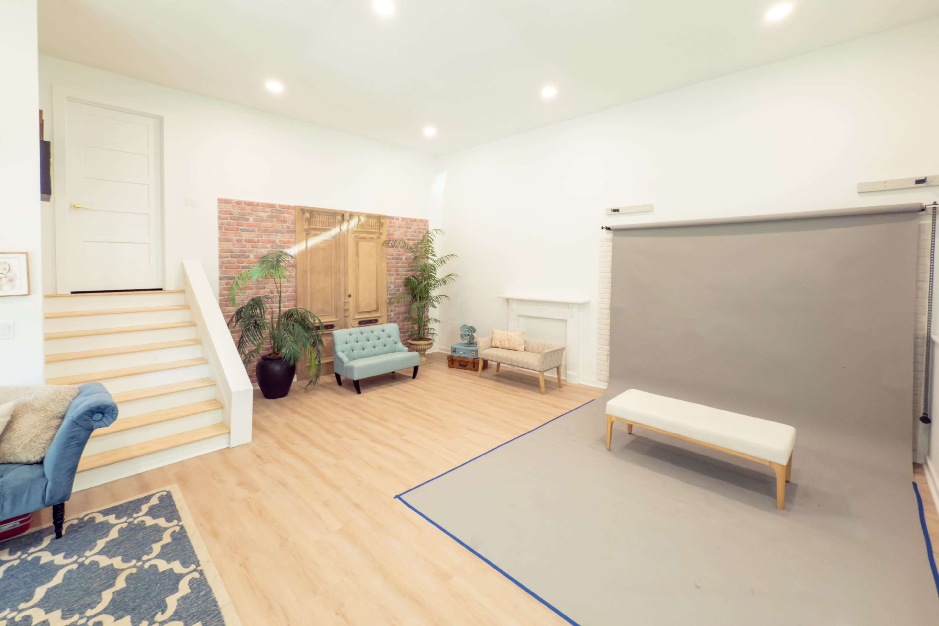 The image shows a bright, spacious room with wooden flooring, a staircase on the left, a grey photography backdrop on the right, and a mix of furniture including a blue couch and a small bench.