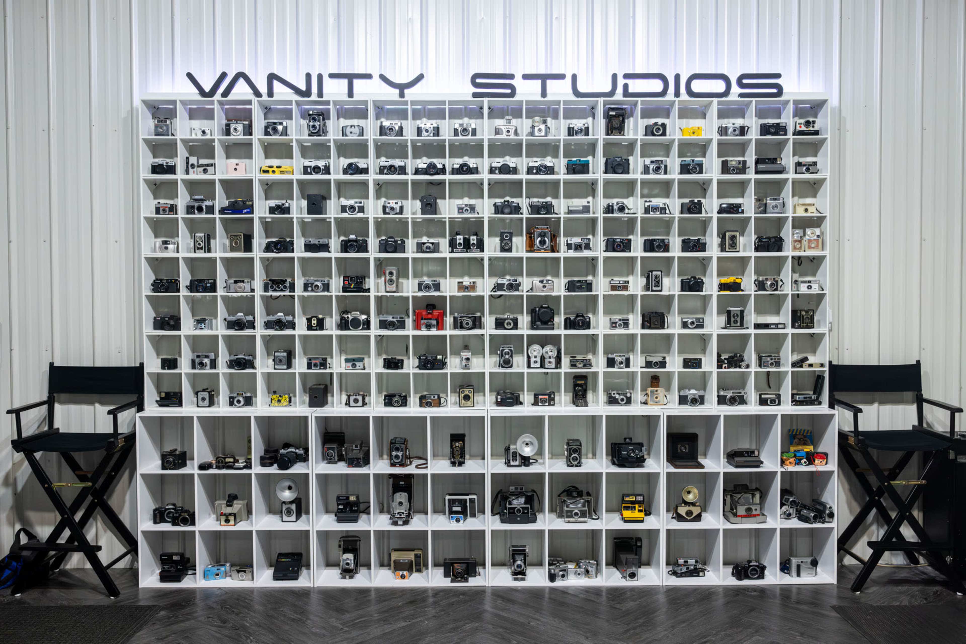 A neatly arranged wall shelf displaying a large collection of cameras and photography equipment, with two director's chairs positioned beside it.