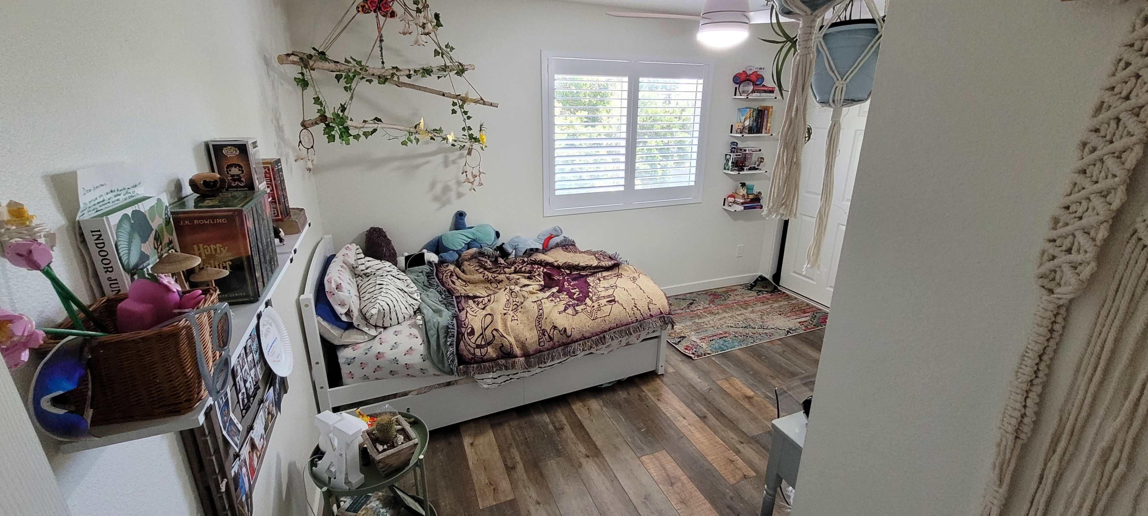 A small bedroom features a bed with a colorful blanket, decorative plants on the walls, and shelves filled with books and toys.