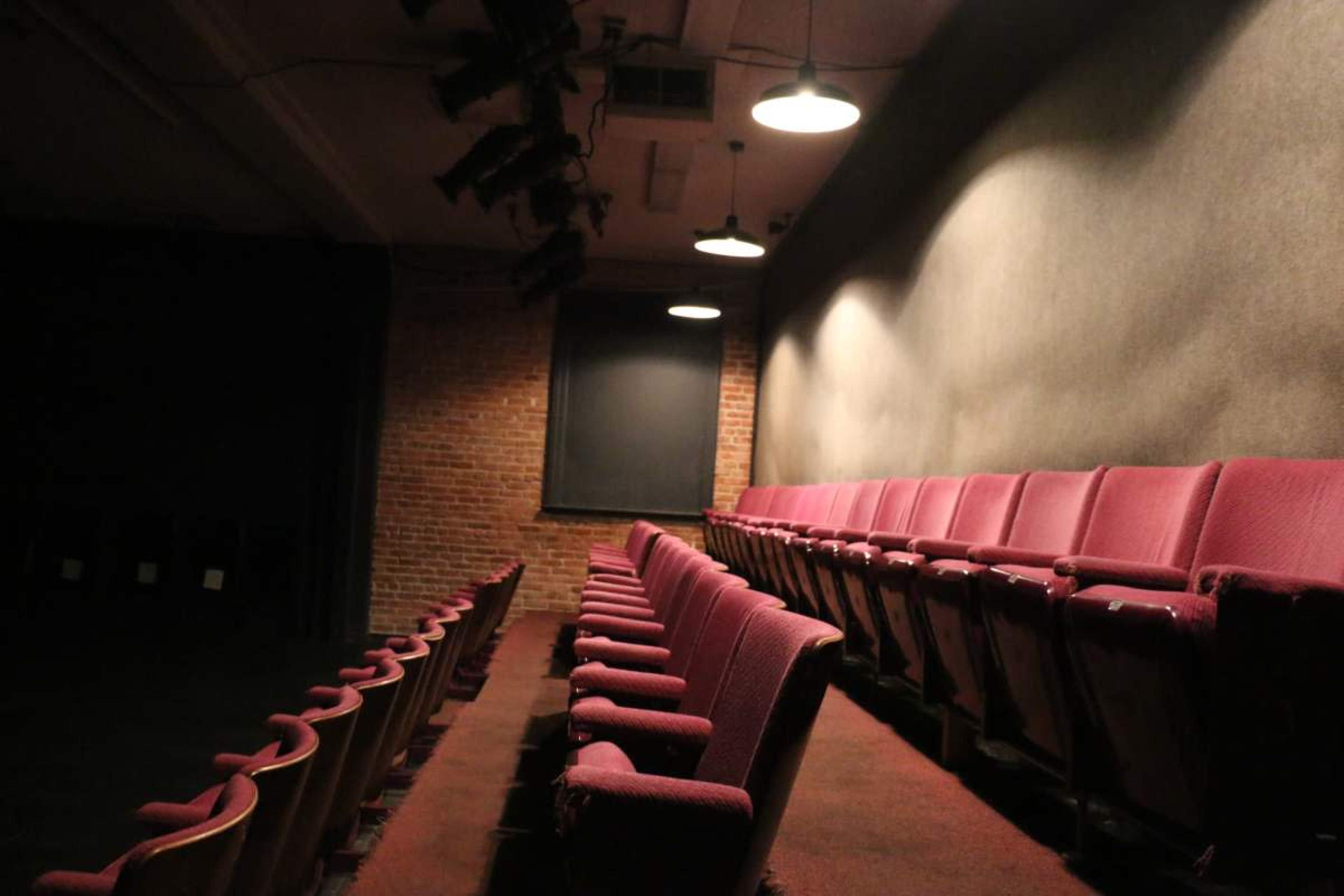 The image shows a dimly lit theater with maroon seating arranged in rows, alongside a brick wall and a large window covered with a dark shade.