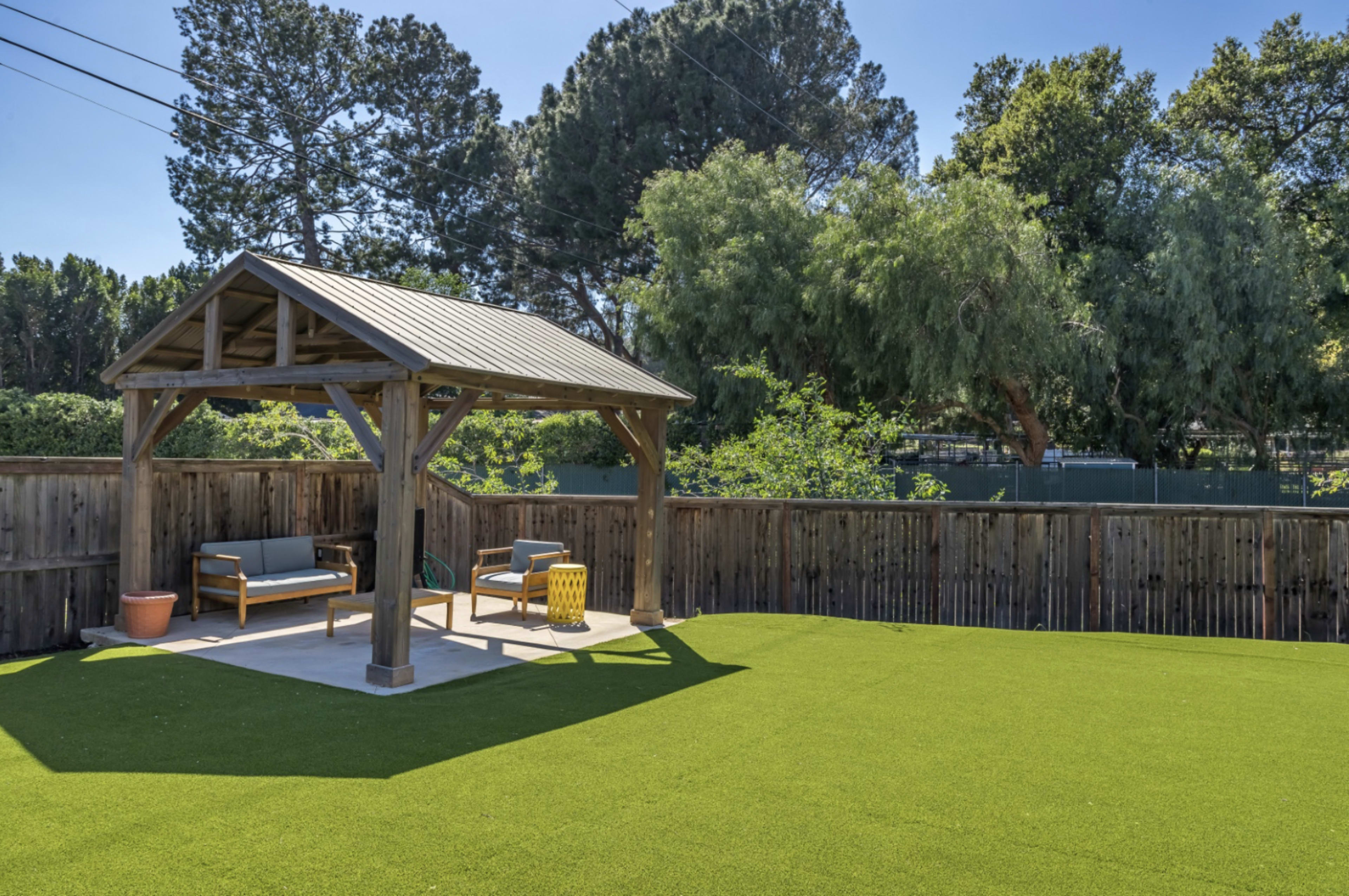 A shaded gazebo with seating is situated on a lawn of artificial turf surrounded by wooden fencing and trees.
