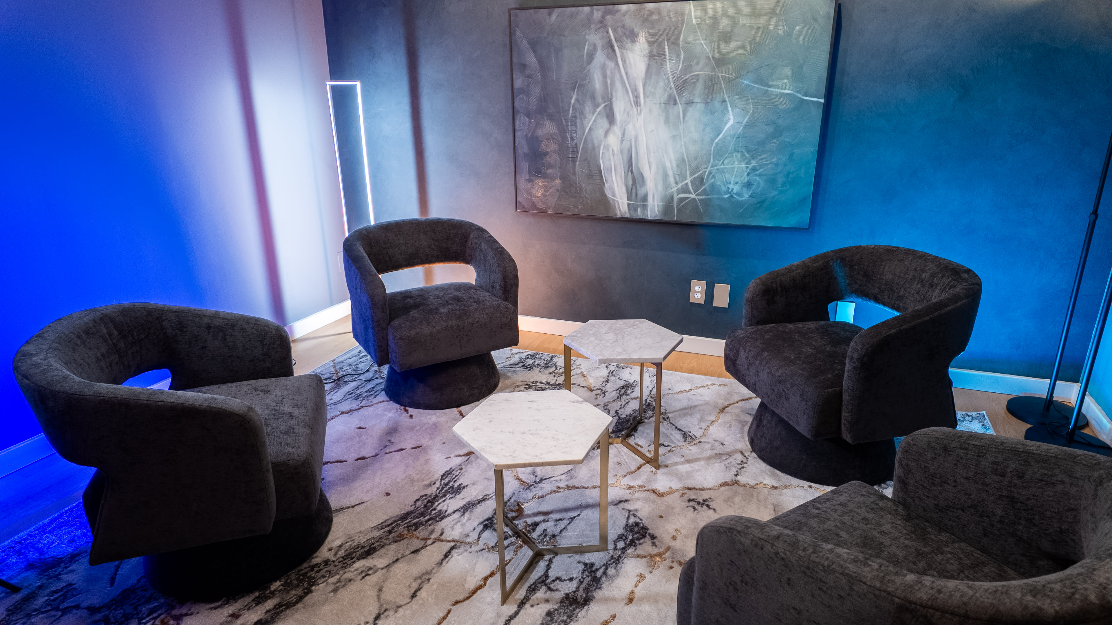 The image shows a modern seating area with four upholstered chairs arranged around two small hexagonal tables on a marbled rug, with abstract artwork hanging on the wall.