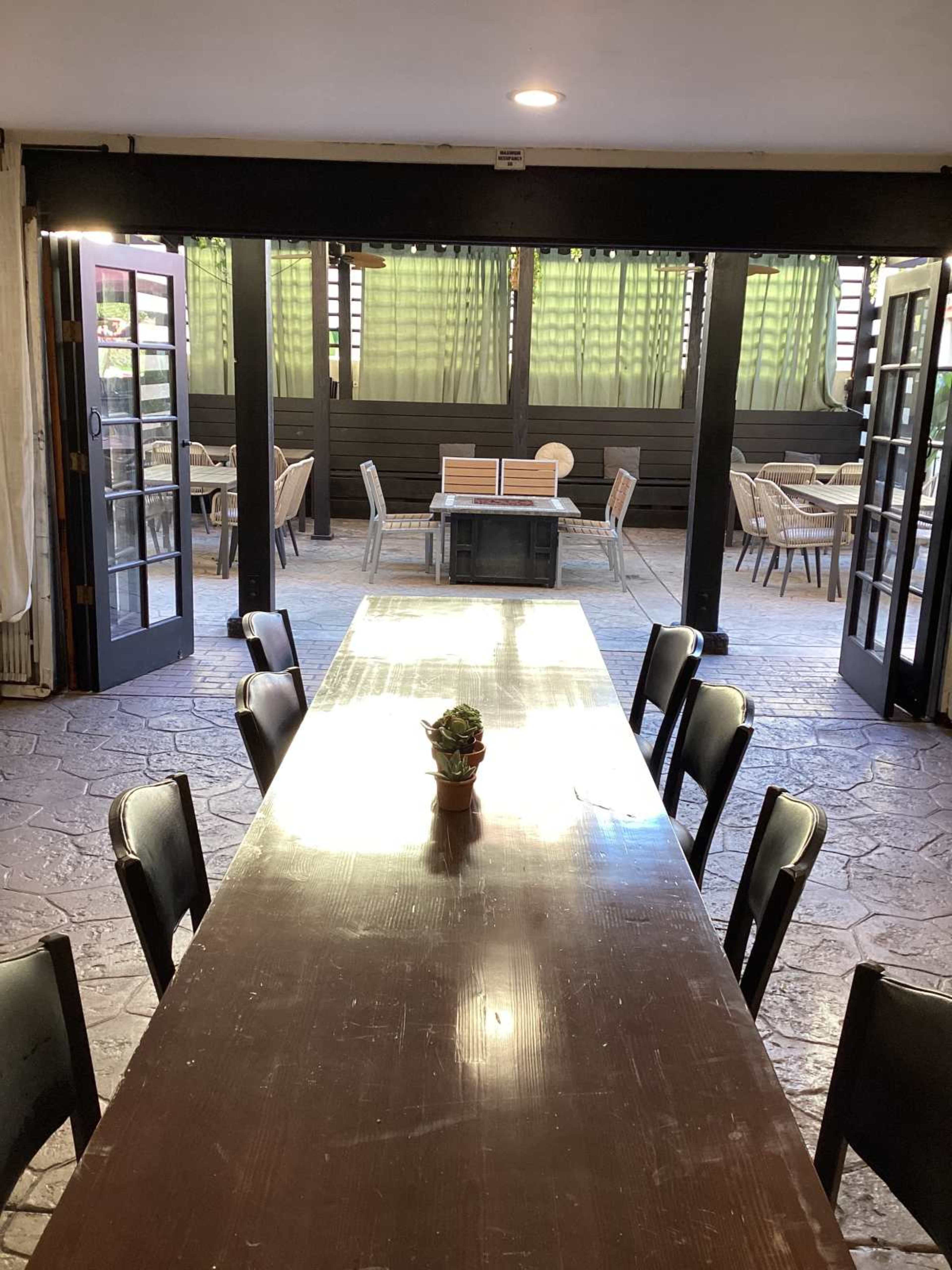 A long wooden table with chairs is set in a dining area, leading to an outdoor space with green curtains and additional seating.