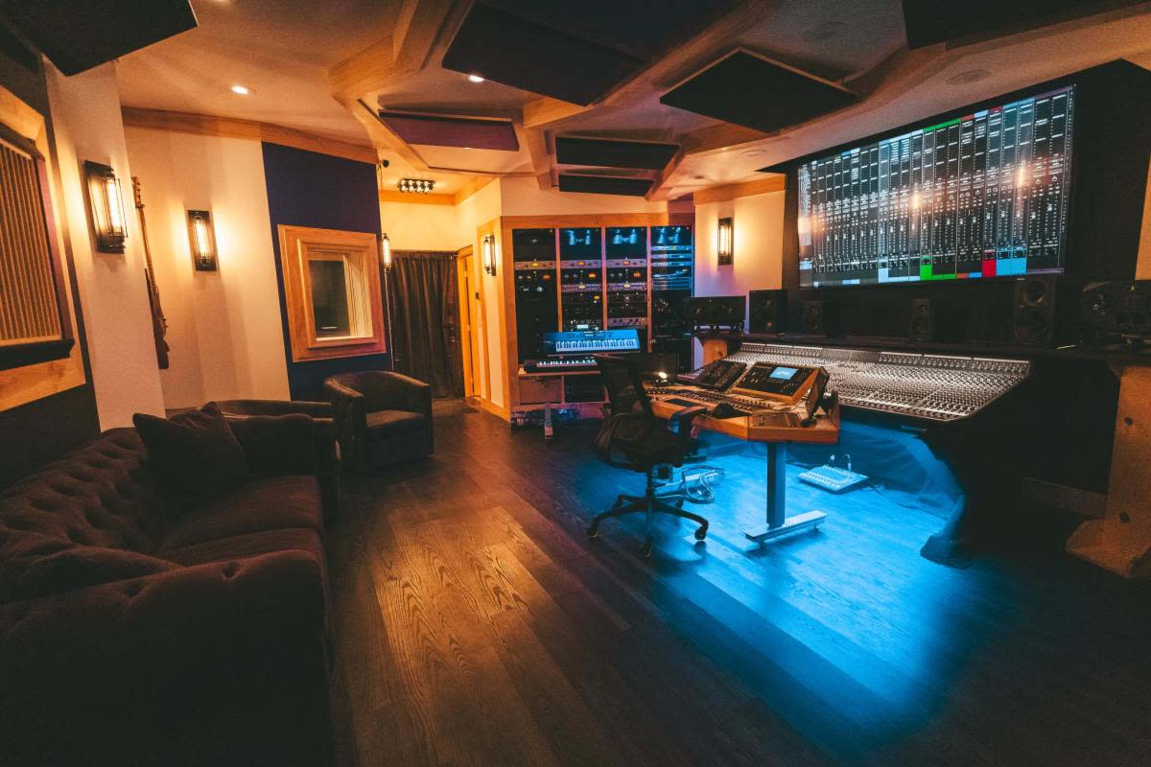 The image shows a well-equipped recording studio featuring a large mixing console, multiple monitors, and a cozy seating area with a dark couch.