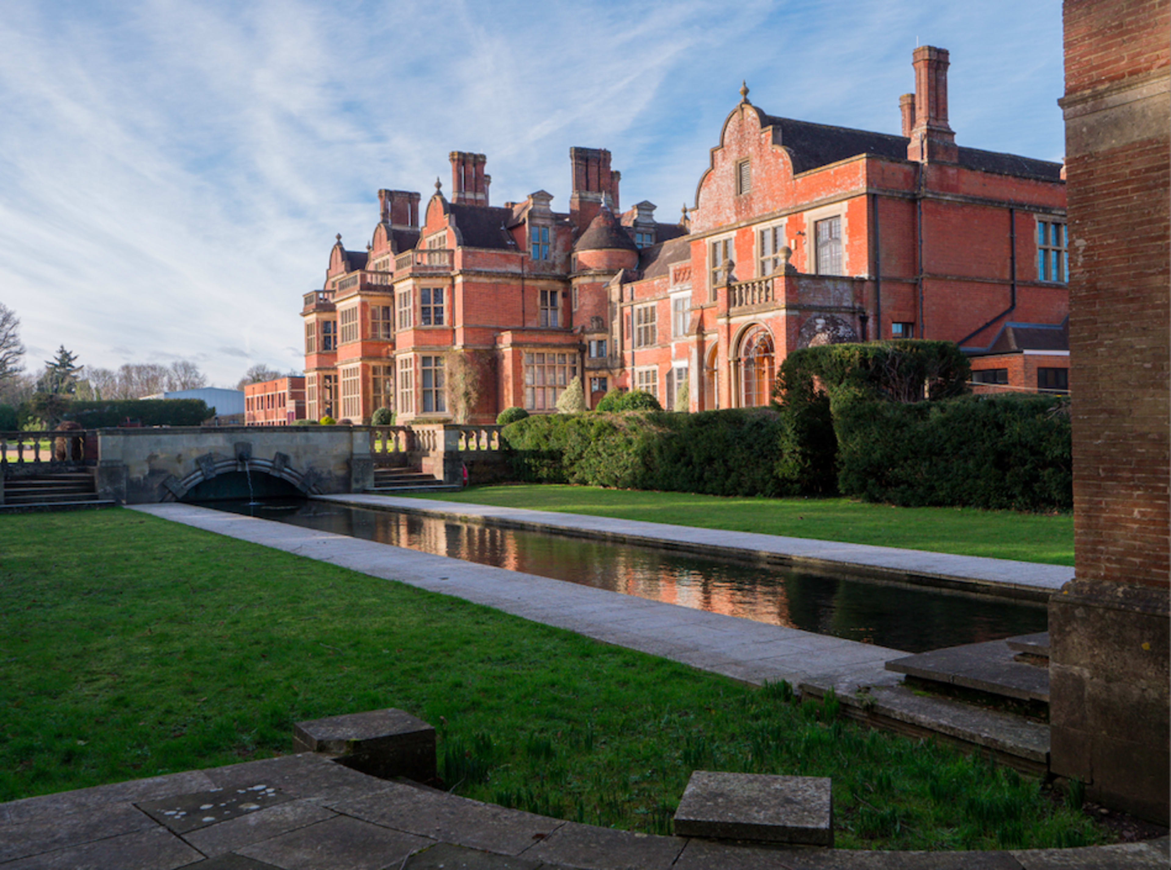 A large red-brick mansion surrounded by neatly trimmed grass and a reflecting water channel in the foreground.