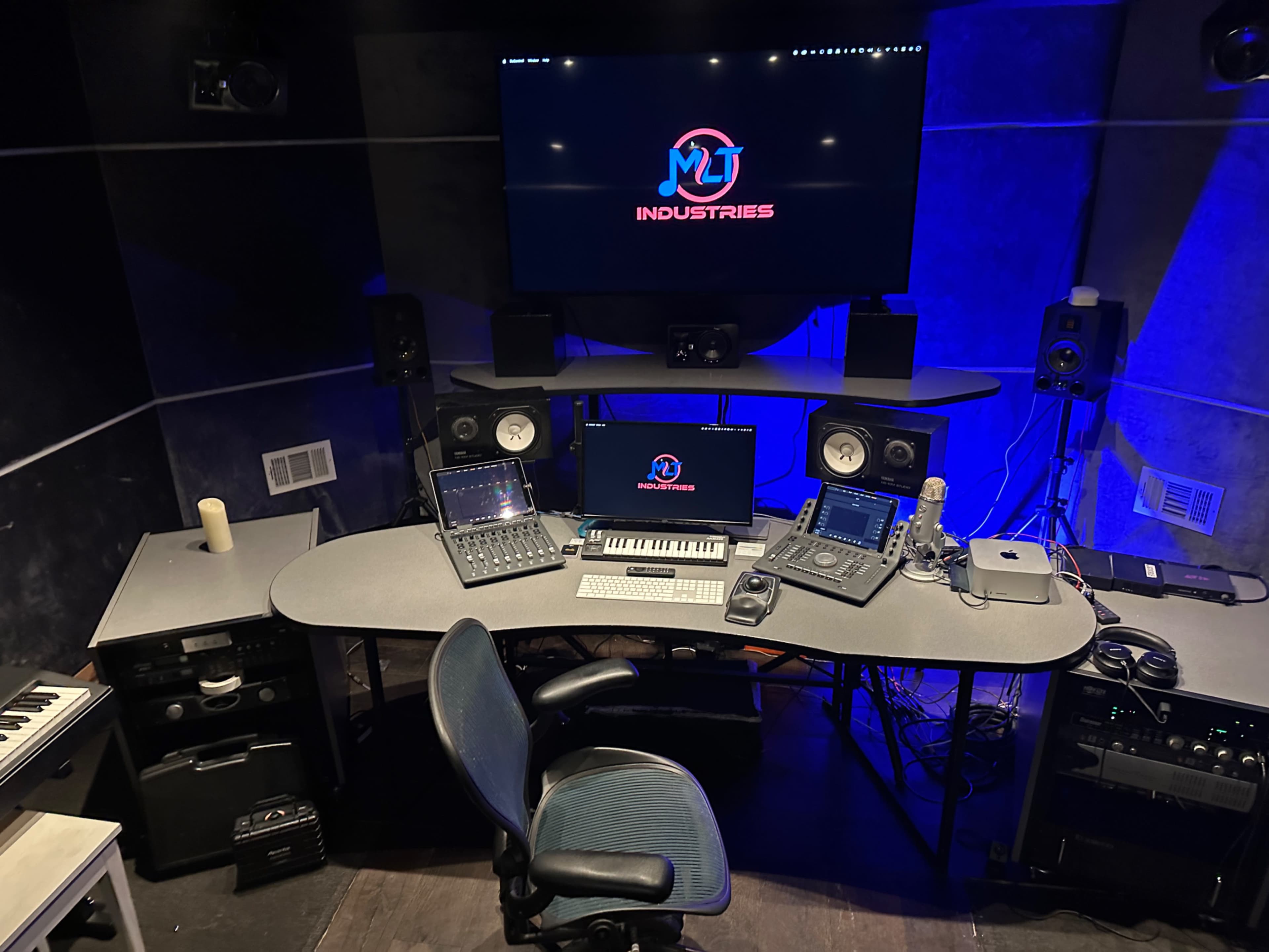 A music production studio features a large screen displaying "MT Industries" and various audio equipment, including a keyboard, mixers, and speakers, arranged around a central desk with a rolling chair.