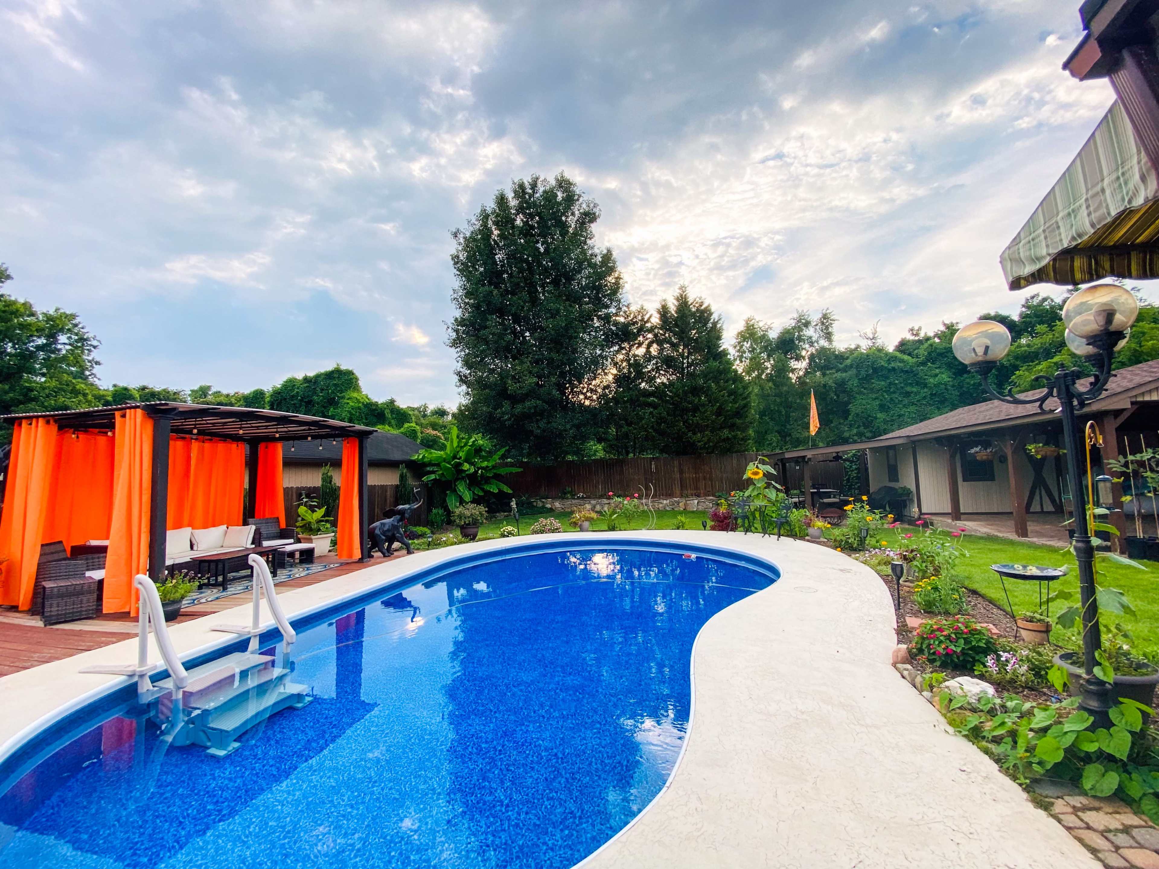The image shows a backyard pool surrounded by greenery, a wooden pavilion with orange curtains, and a garden area with flowers and decorations.