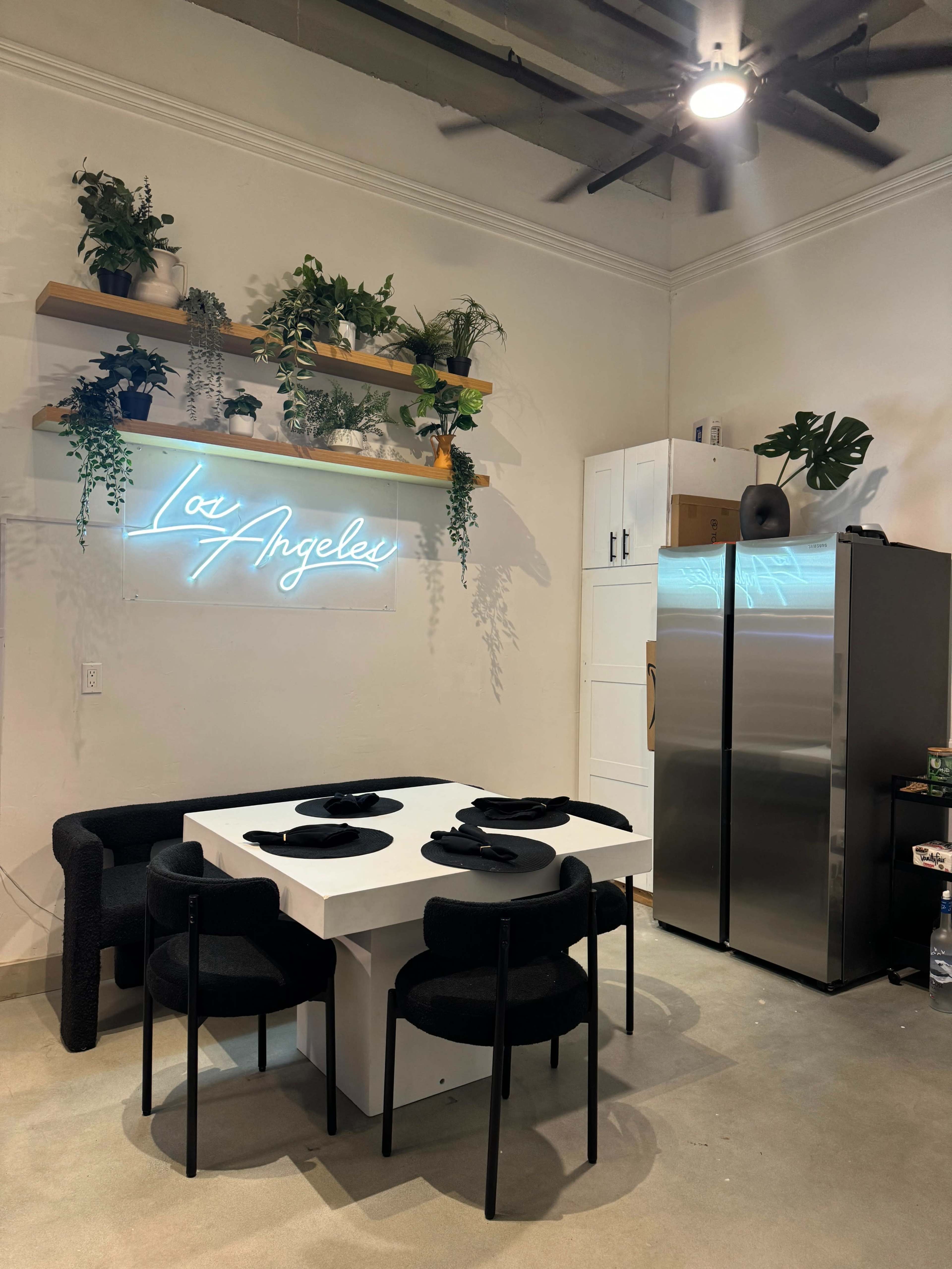 The image shows a modern kitchen area featuring a white table with four black chairs, a stainless steel refrigerator, and shelves adorned with various plants, illuminated by a neon sign that reads "Los Angeles."