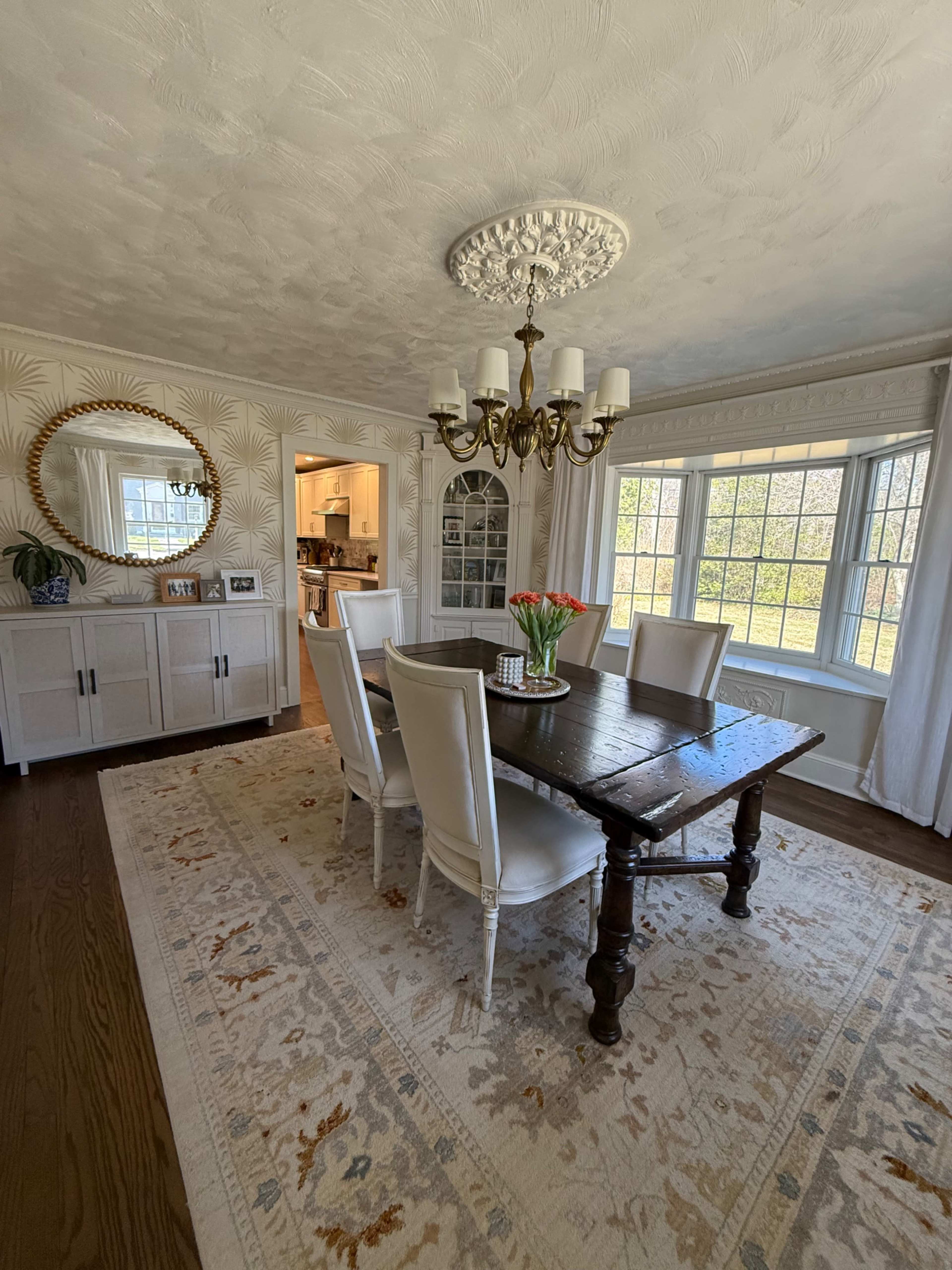 The image shows a well-lit dining room with a wooden table, five white upholstered chairs, a chandelier, and a large mirror reflecting the space.