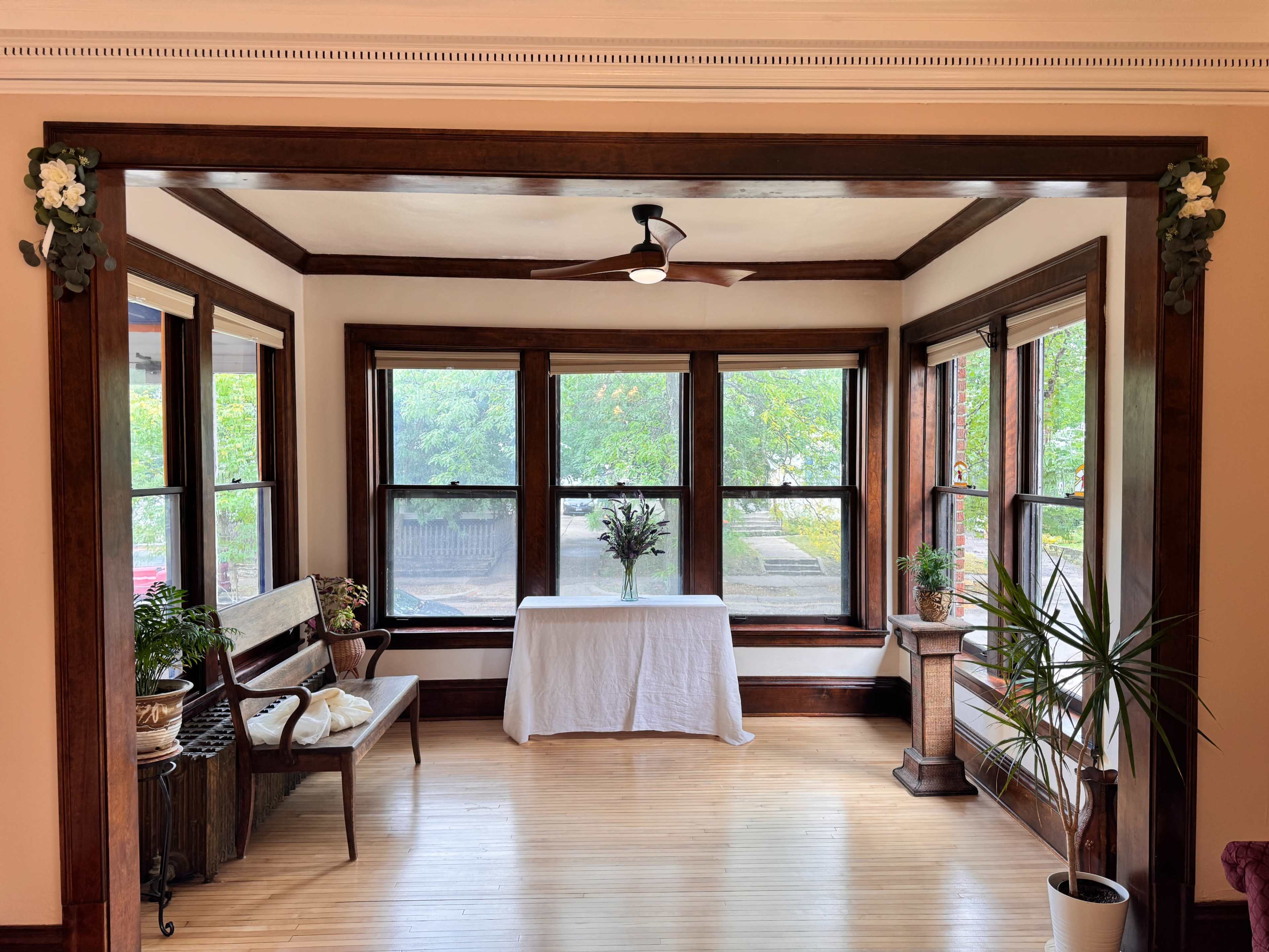 The image shows a sunlit room featuring large windows, a small table with a floral centerpiece, a bench, and a potted plant.