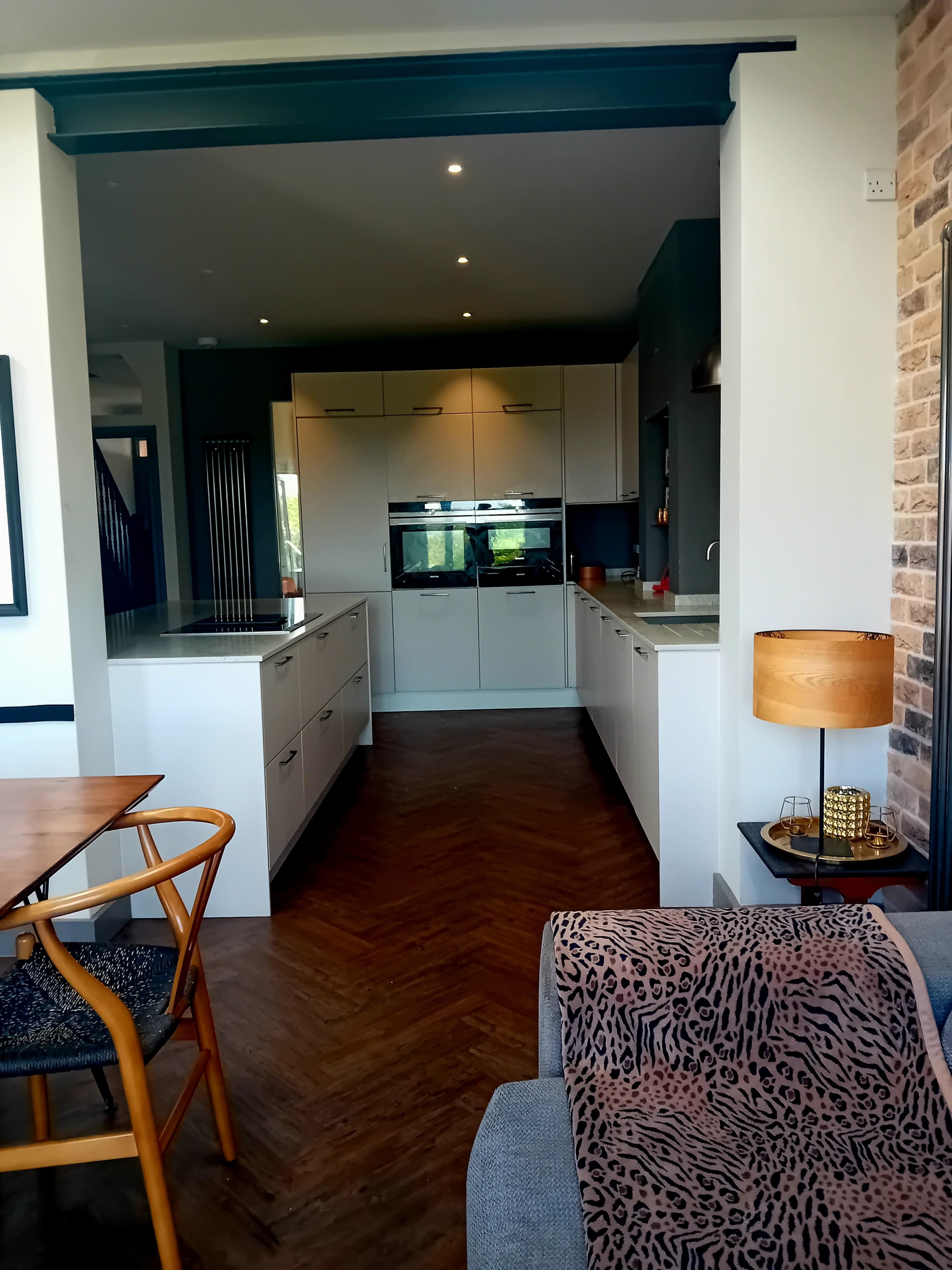 A modern kitchen with white cabinets and a herringbone-patterned wood floor connects to a living area featuring a table and a patterned throw on a gray couch.