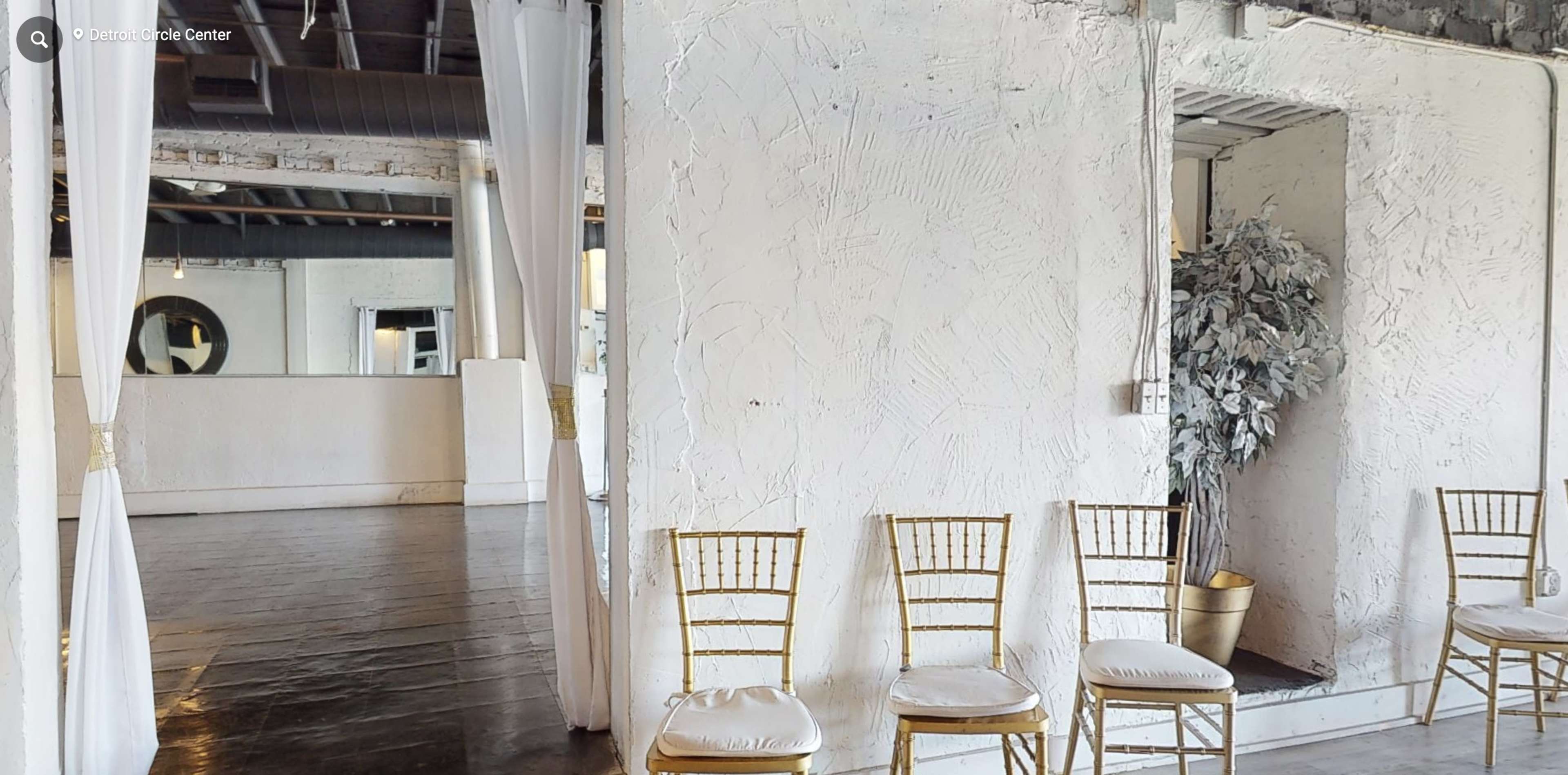 A spacious, well-lit room with empty gold chiavari chairs lined up against a textured white wall.