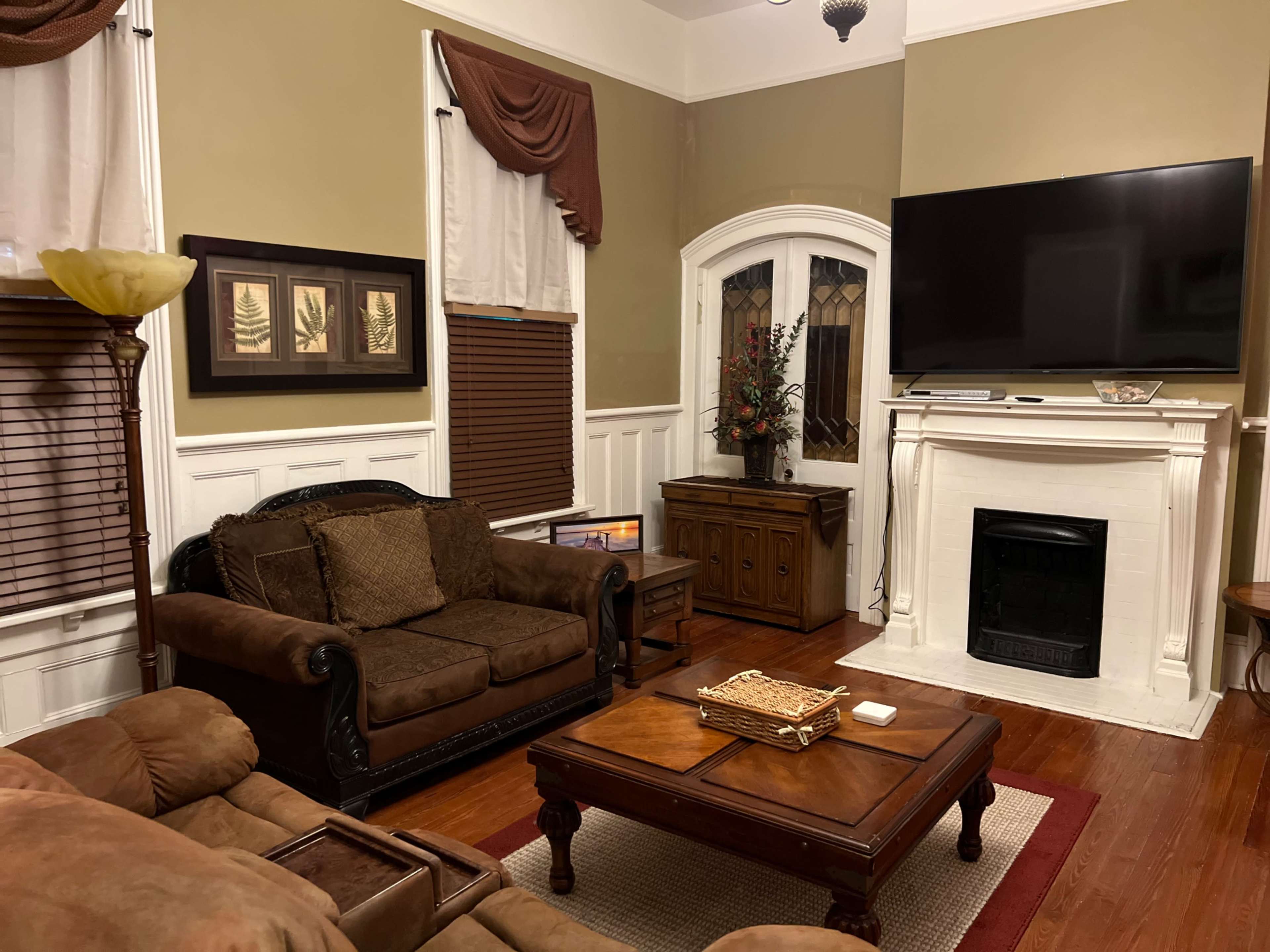 The living room features a dark brown couch and loveseat, a wooden coffee table, and a mounted television above a white fireplace, with decorative elements on the walls and window treatments.
