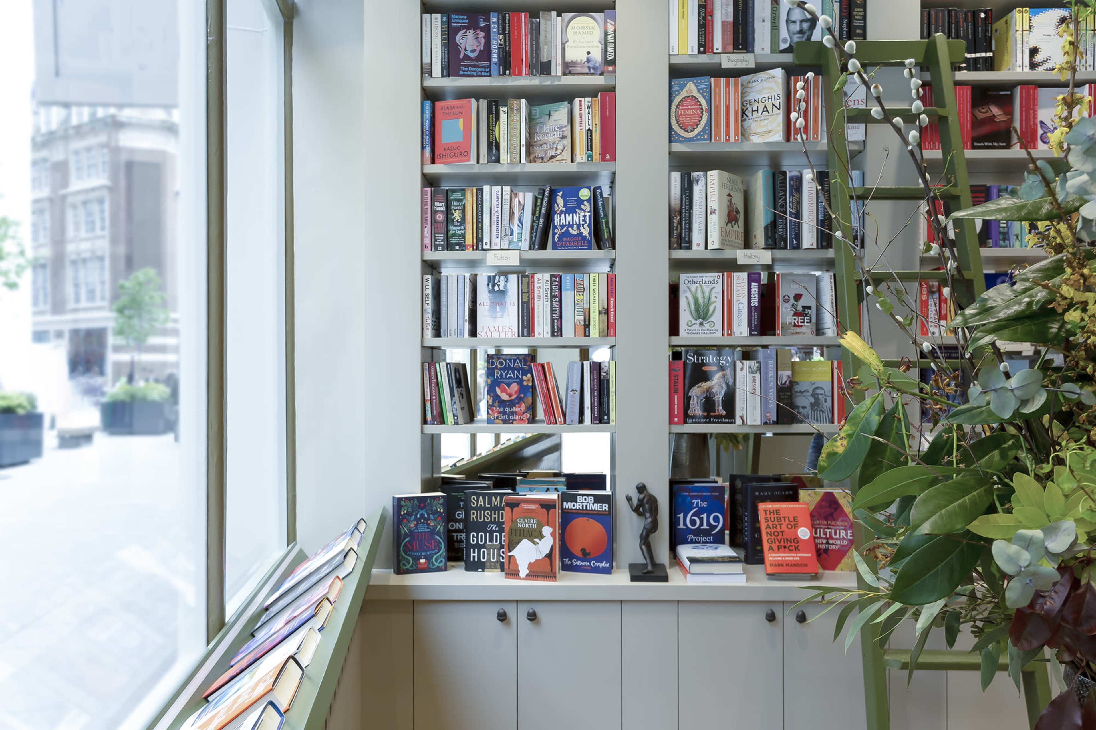 A bookshelf filled with various books is displayed against a wall in a well-lit corner of a modern bookstore, with a table and decorative plants in the foreground.
