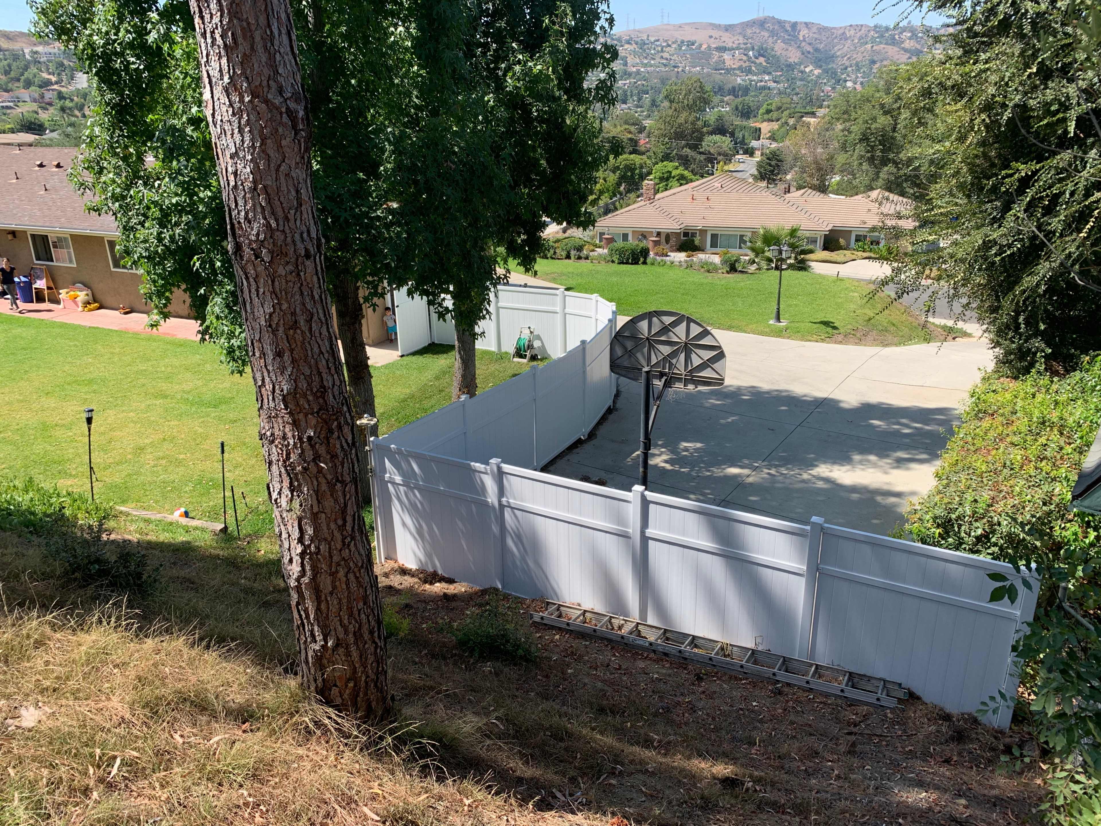 The image shows a sloped backyard with a white fence, a basketball hoop on a concrete surface, and several houses in the background surrounded by greenery.