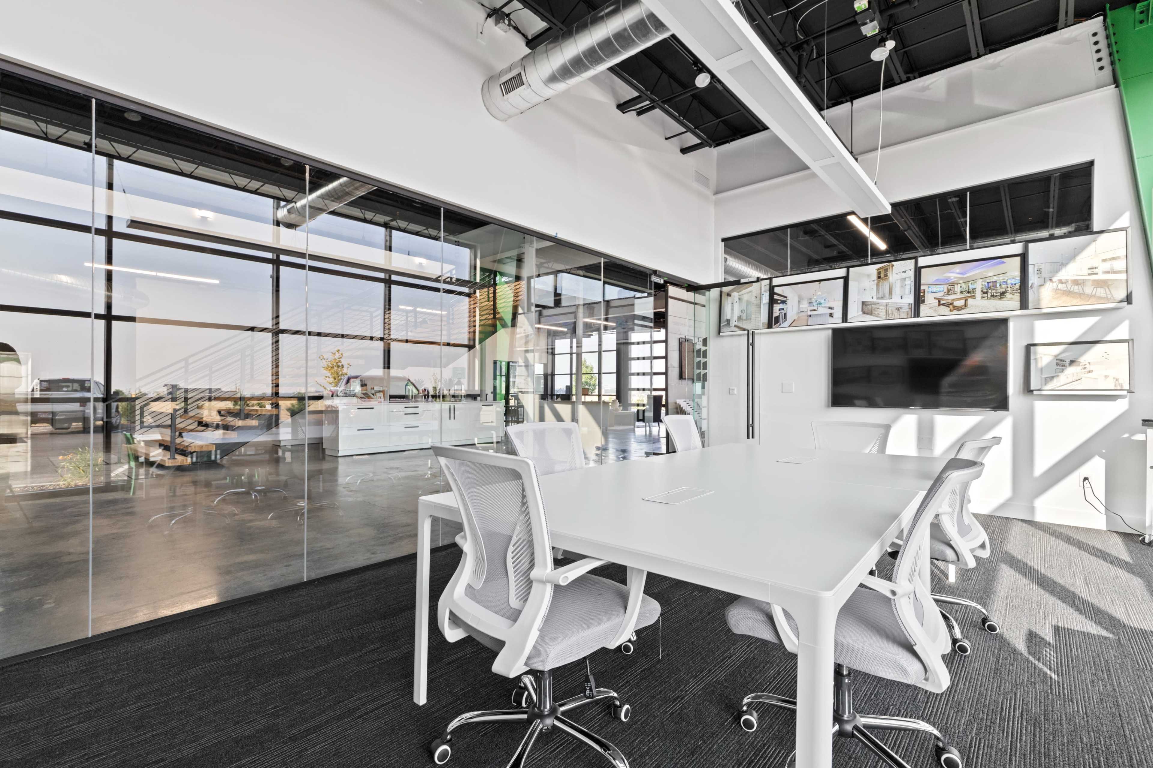 A bright conference room features a large white table surrounded by modern chairs, with glass walls that provide a view of a spacious office area.