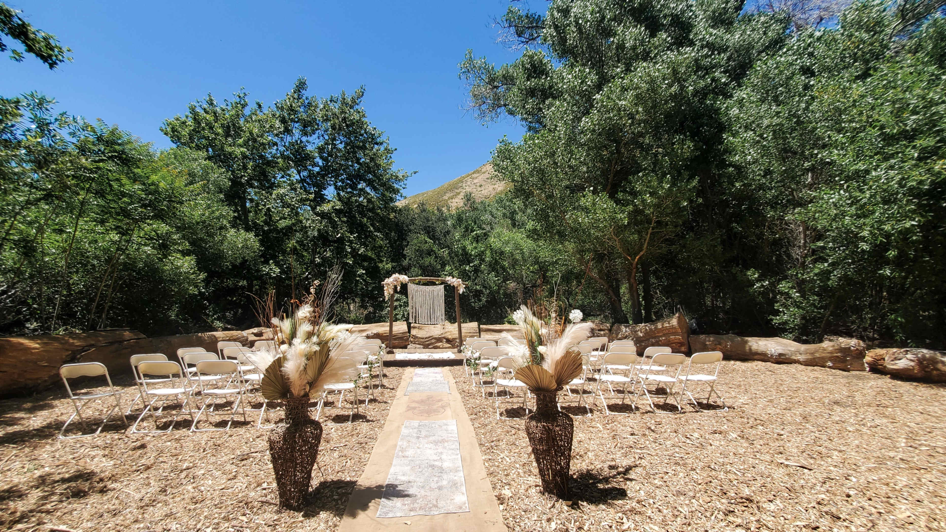 A serene outdoor wedding venue features rows of white chairs arranged on a wooden path, flanked by decorative vases, under a blue sky surrounded by trees.