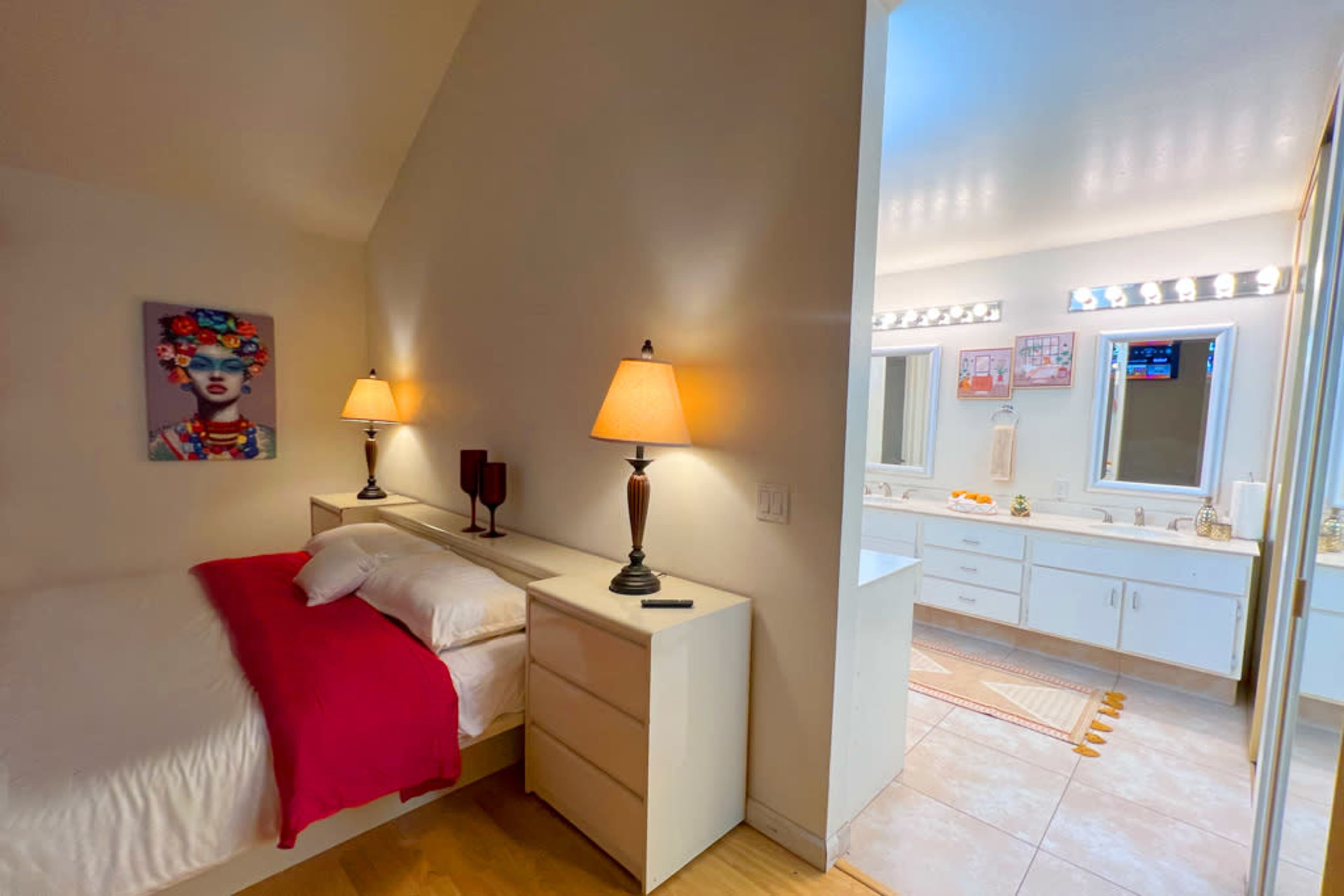 A bedroom with a white bed and red accent, adjacent to a well-lit bathroom featuring a double vanity and large mirrors.