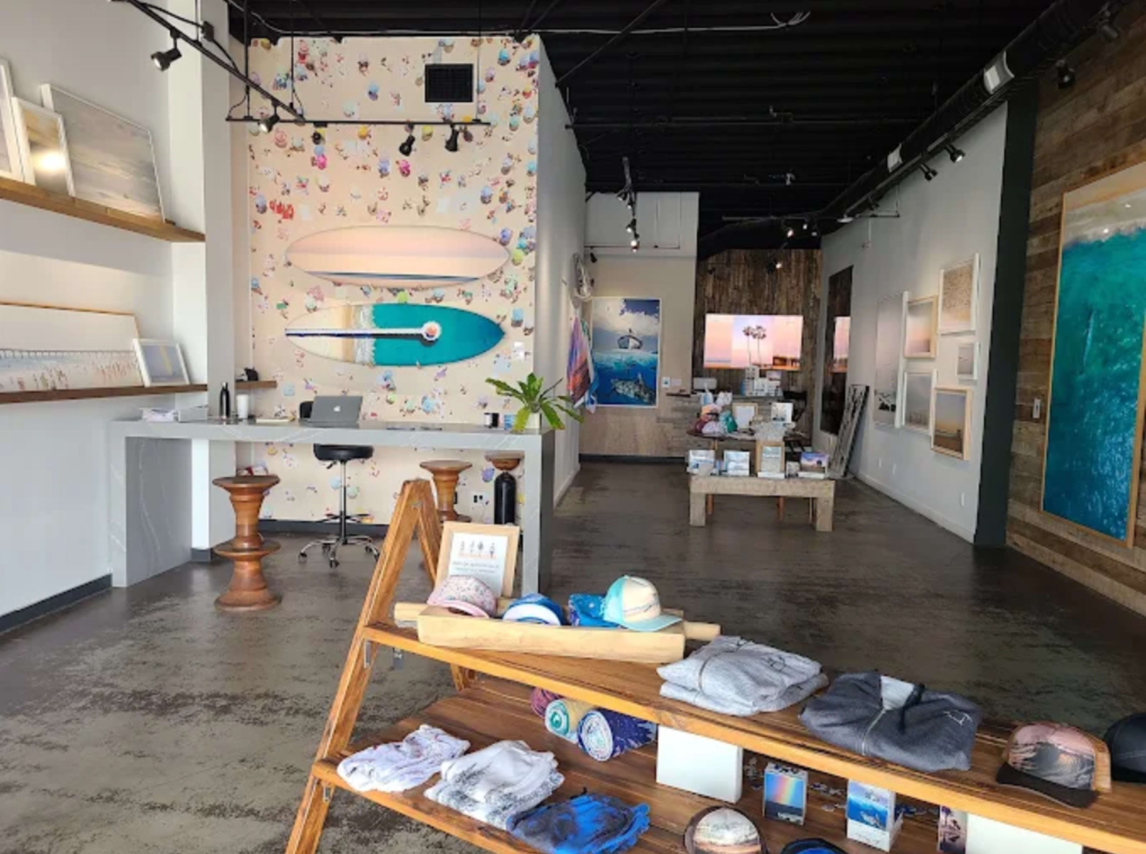 The image shows an interior retail store featuring a display of merchandise on wooden shelves, a reception area with a desk, and decorated walls with artwork and a surfboard motif.