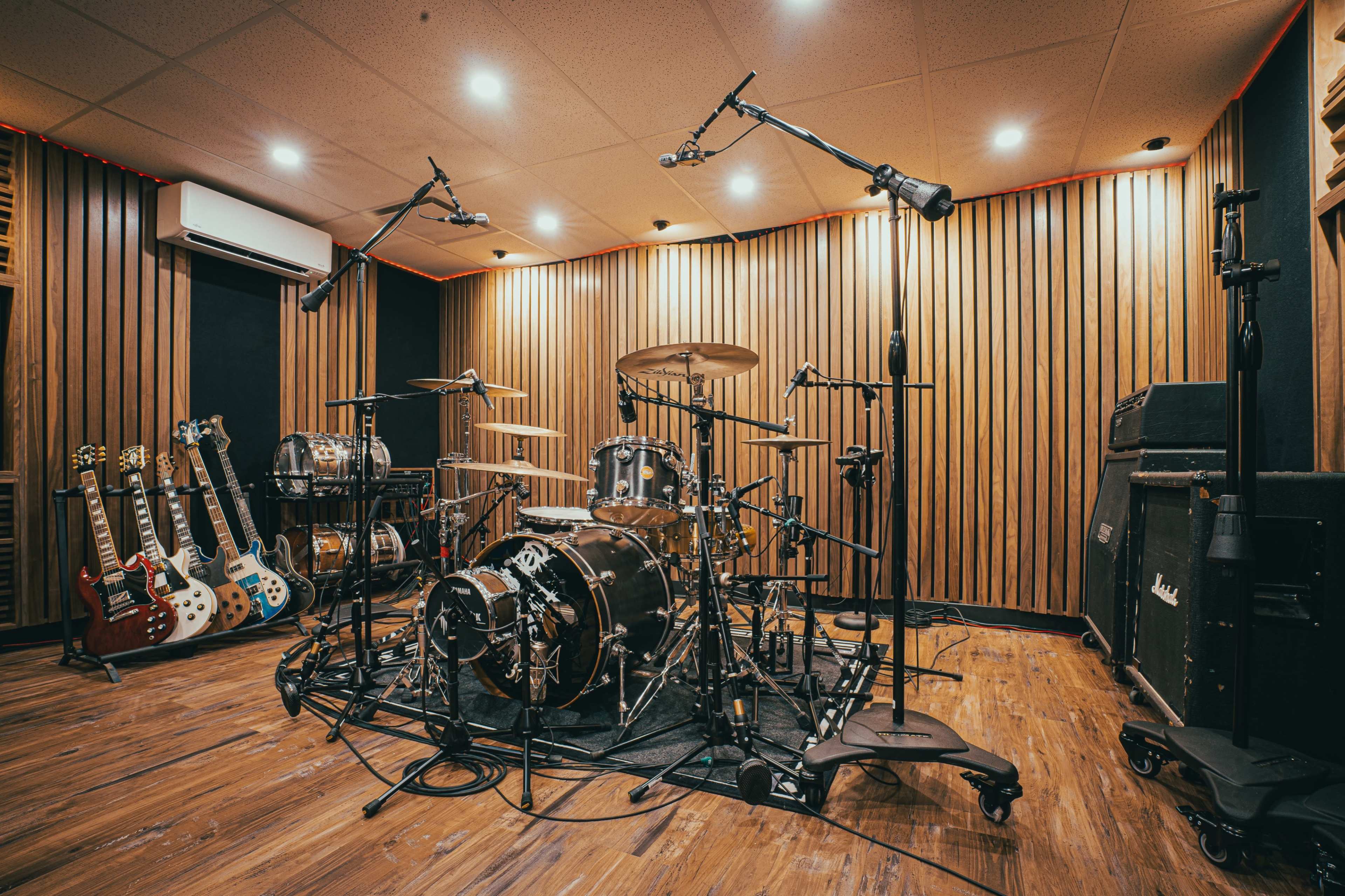 A professional recording studio features a drum set surrounded by multiple microphones, wooden wall panels, and several electric guitars on stands.