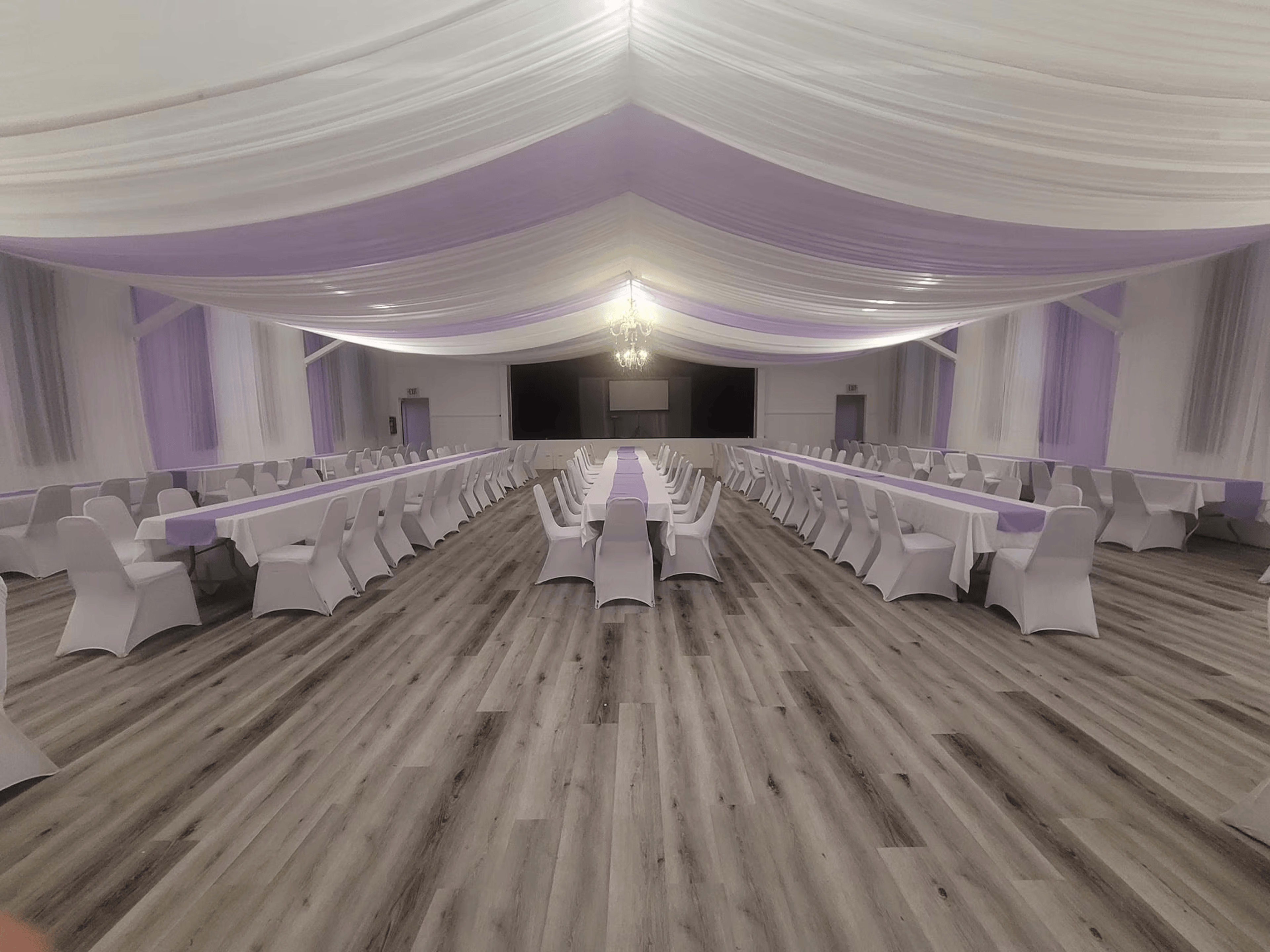 A spacious banquet hall features rows of white-covered chairs and tables arranged under a draped ceiling with purple accents.