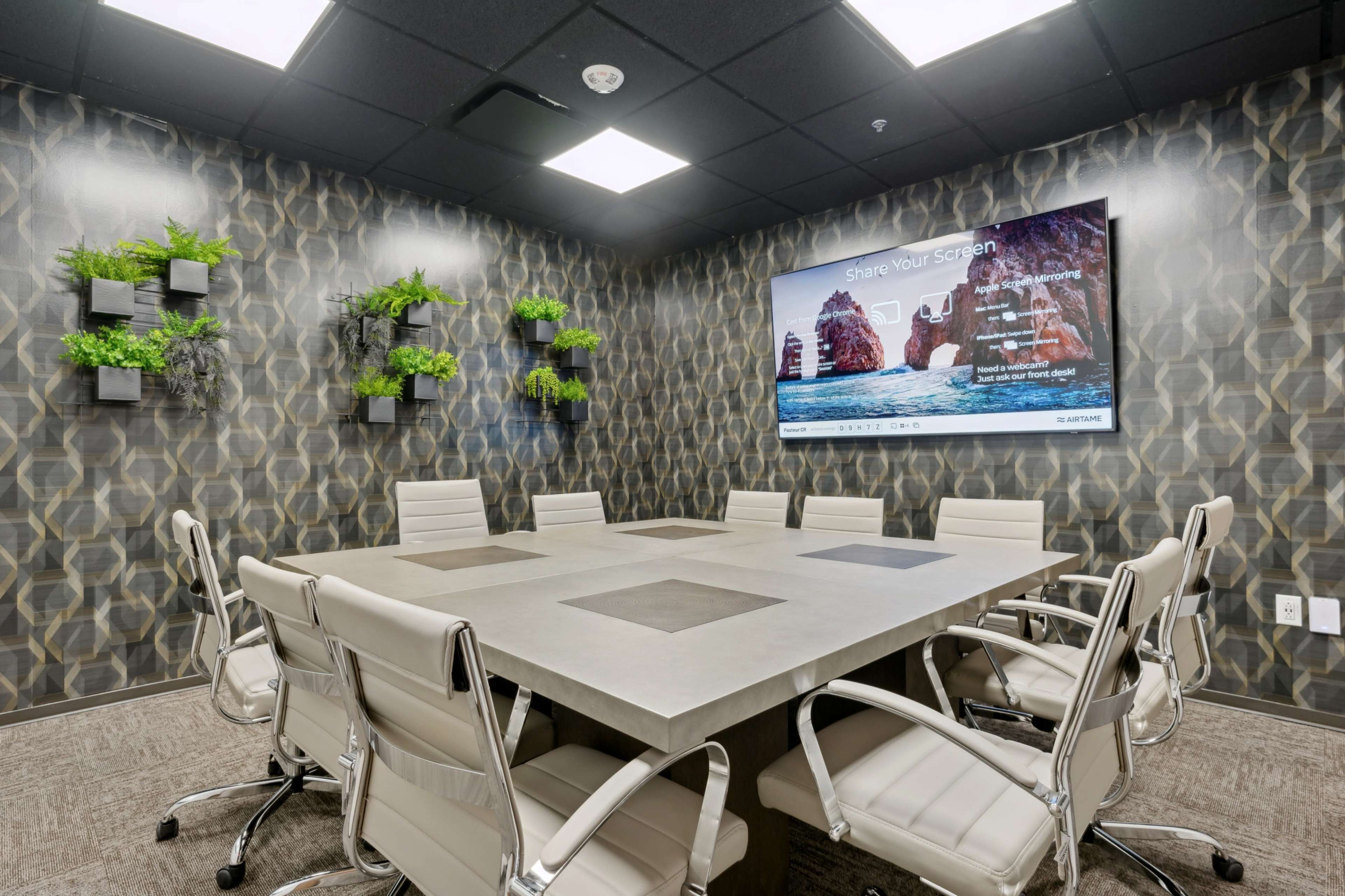 A modern conference room features a large table surrounded by white ergonomic chairs and a screen displaying a presentation on a textured wall adorned with plants.