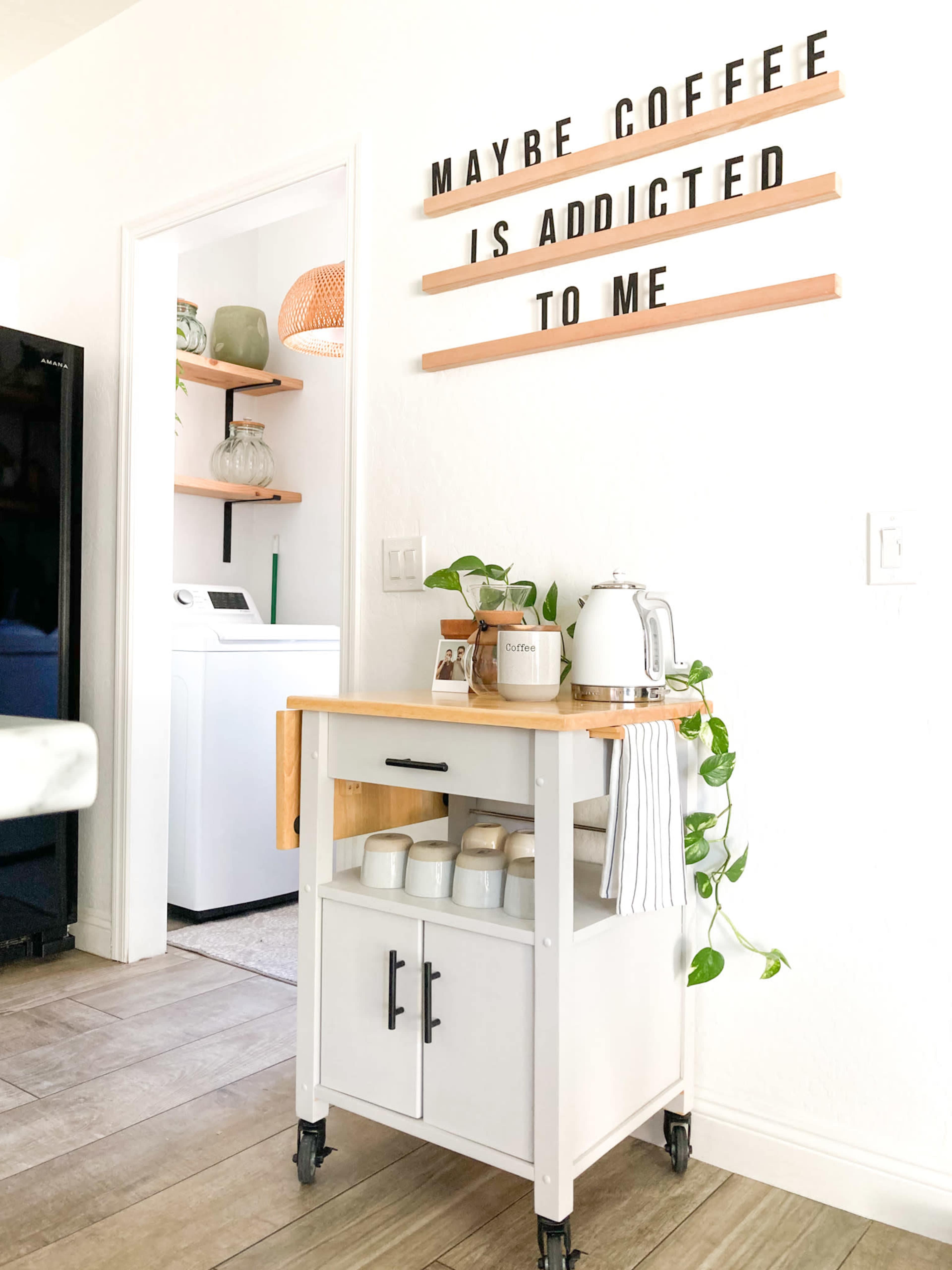 A small kitchen cart with a wood top and white base, adorned with potted plants and a kettle, is positioned near a wall displaying a wooden sign with the words "MAYBE COFFEE IS ADDICTED TO ME."