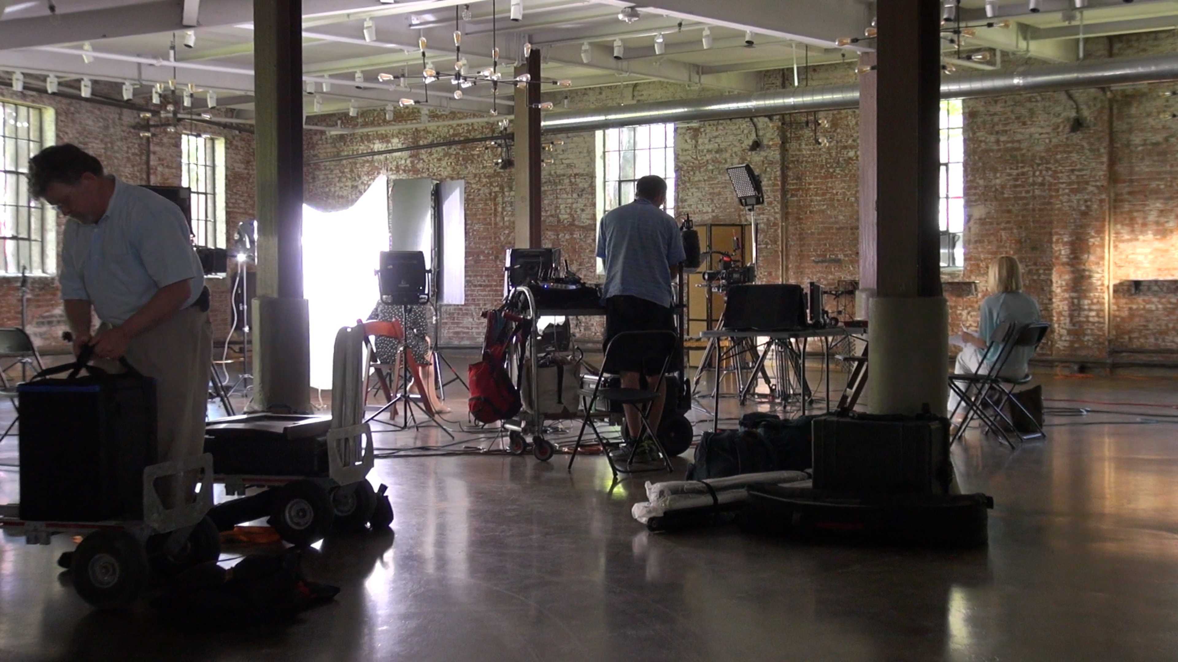 A film production crew sets up equipment in a spacious industrial workspace with large windows and exposed brick walls.