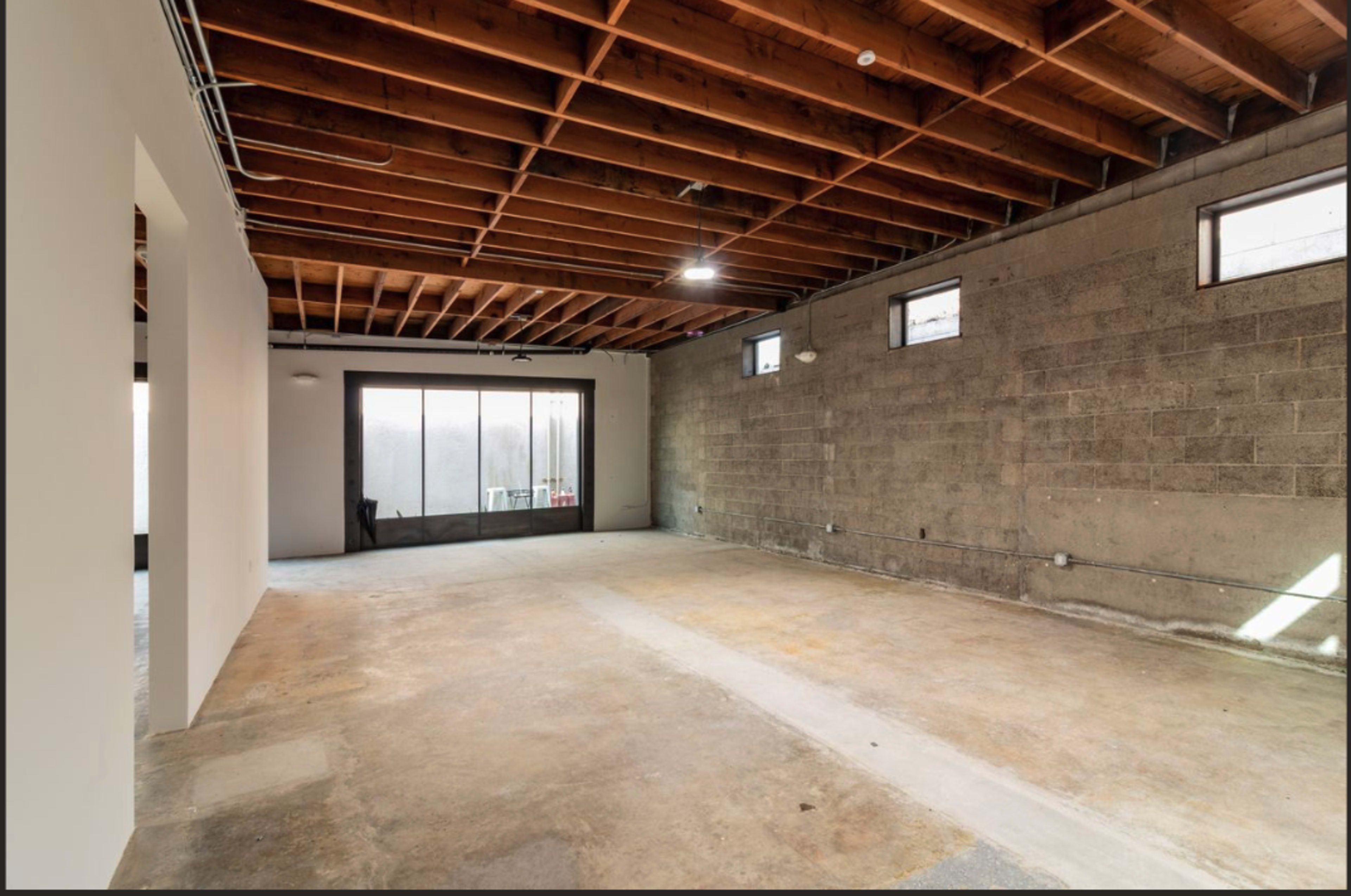 The image shows an empty room with exposed wooden beams on the ceiling and concrete walls, featuring large windows that allow natural light to enter.