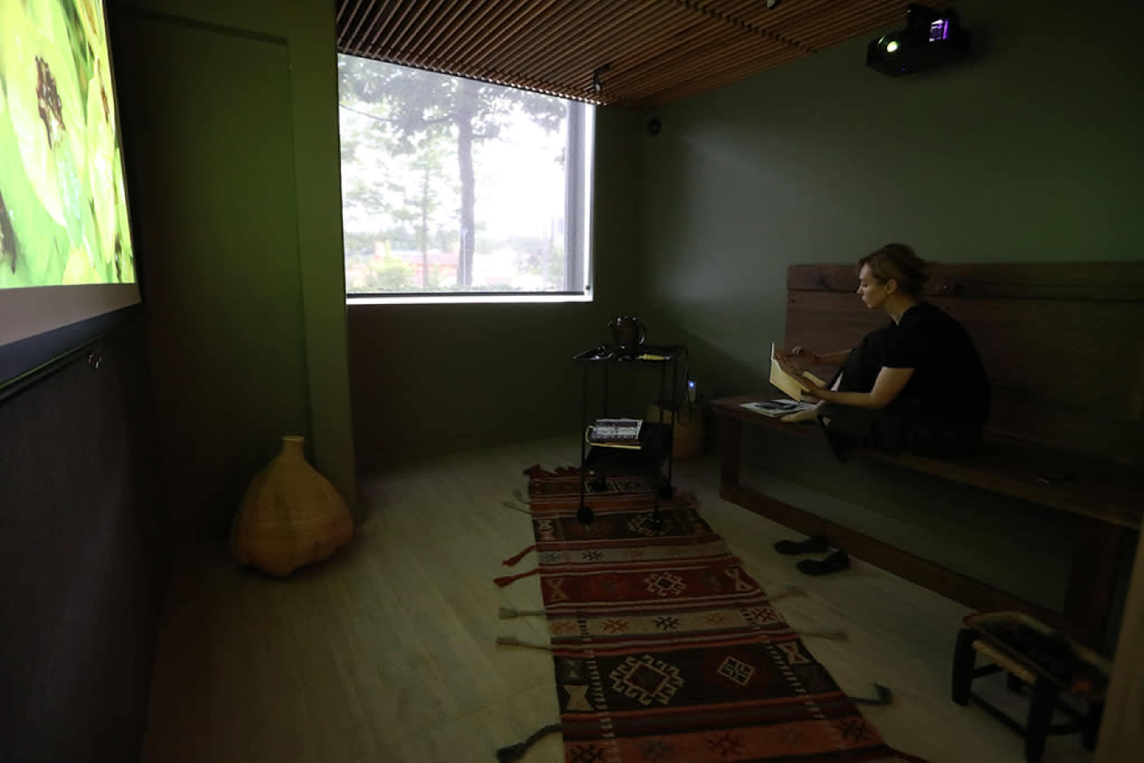 A person sits on a bench in a small, dimly lit room with a projector displaying an image on the wall.