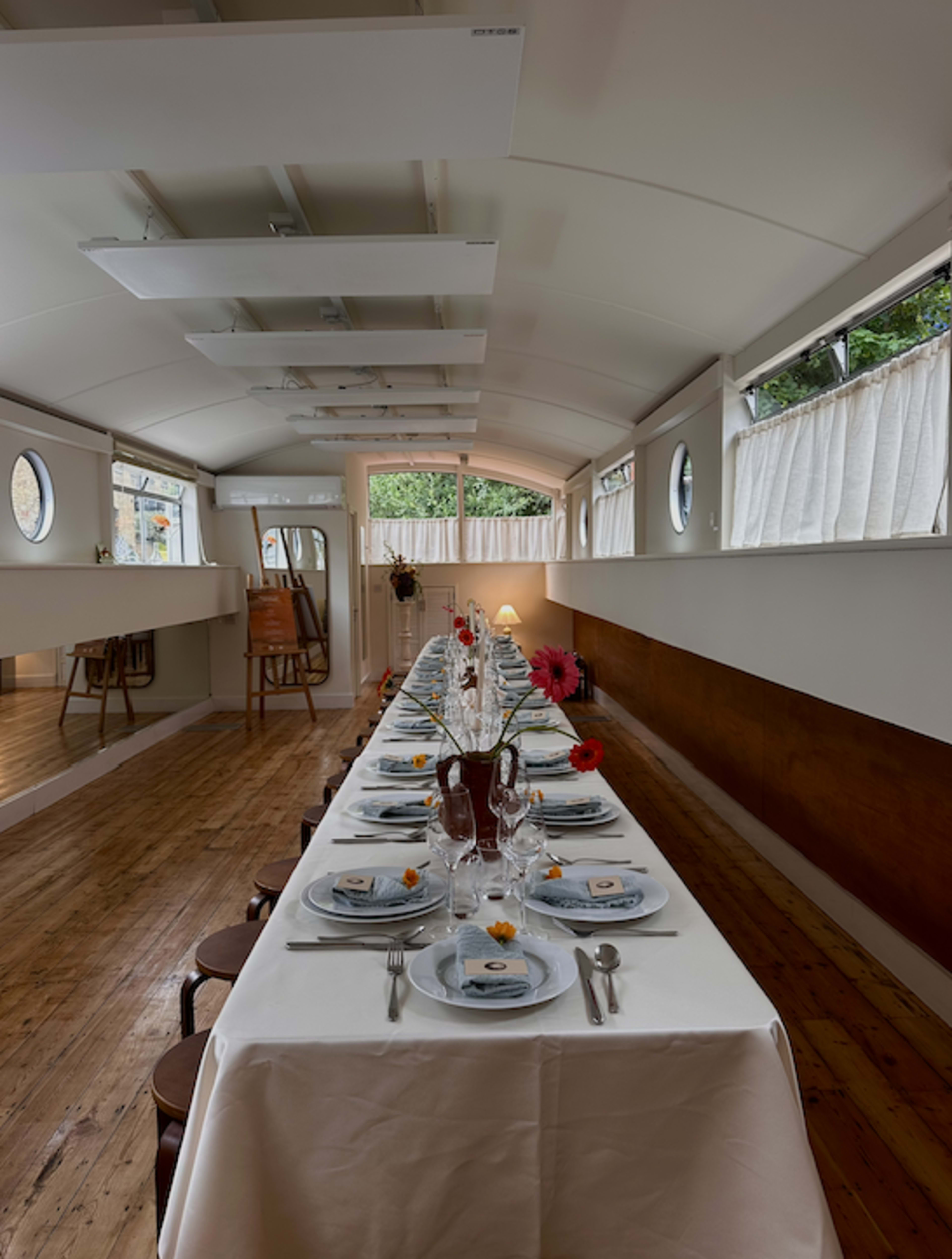 A long dining table is set with plates and glasses inside a brightly lit, modern dining space featuring large windows and a wooden floor.