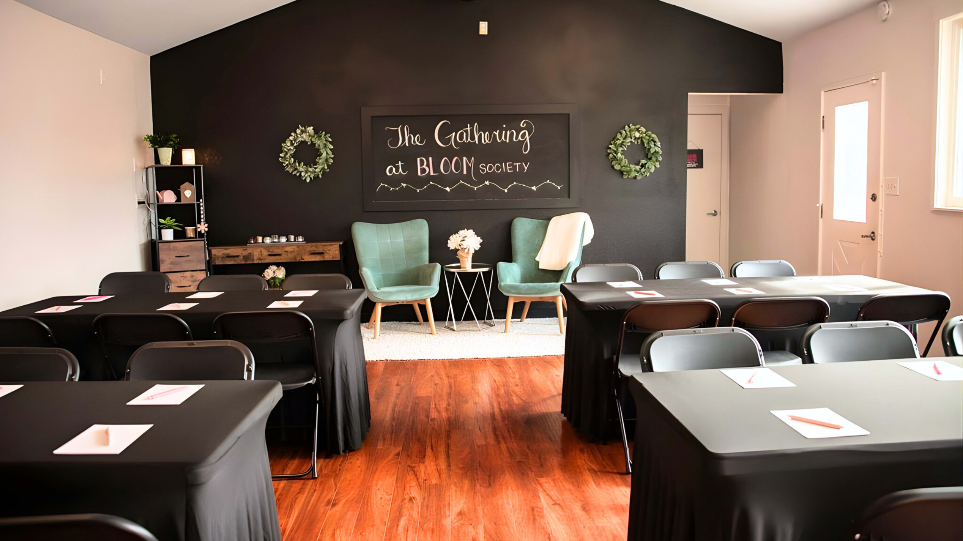 The image shows a meeting room with several tables covered in black tablecloths, green chairs, and a black wall featuring decorative wreaths and a chalkboard sign.