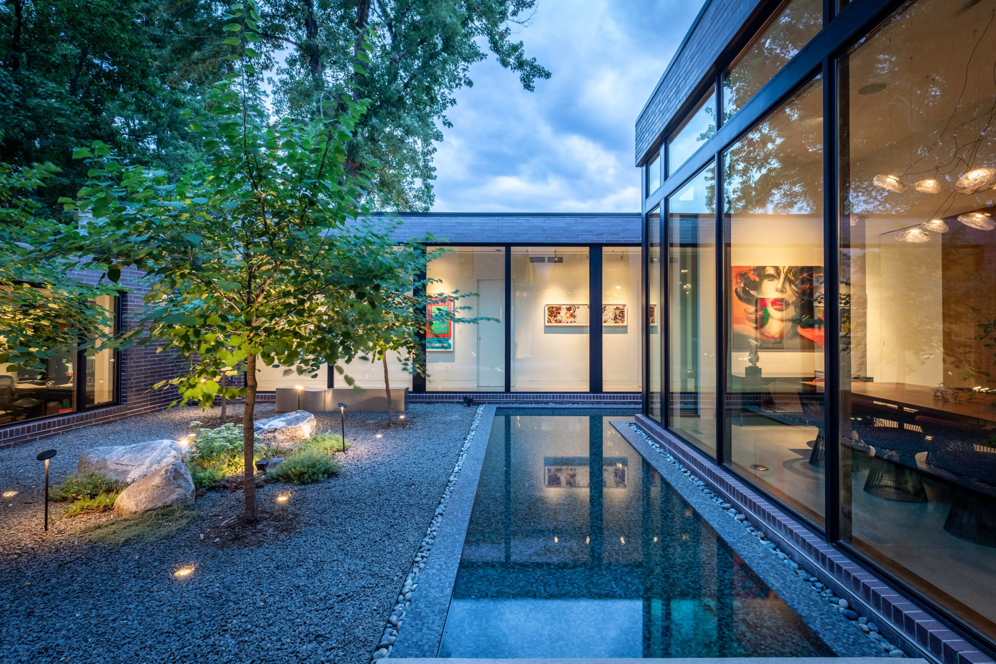 The image shows a modern house with large glass windows, surrounded by a landscaped garden featuring boulders, gravel, and a reflecting pool.