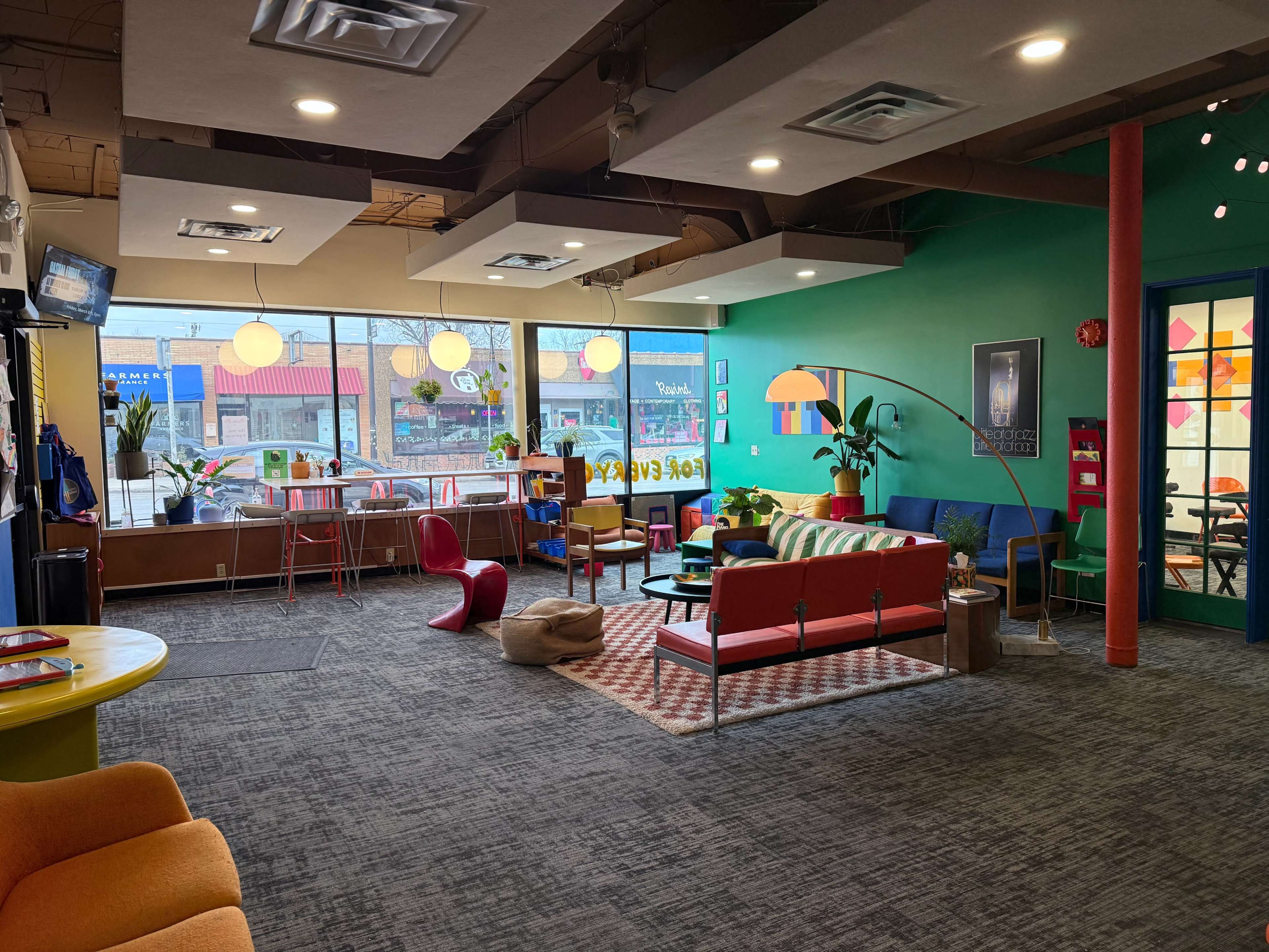 The image shows a colorful, modern lounge area with various seating options, plants, and large windows displaying an exterior street view.