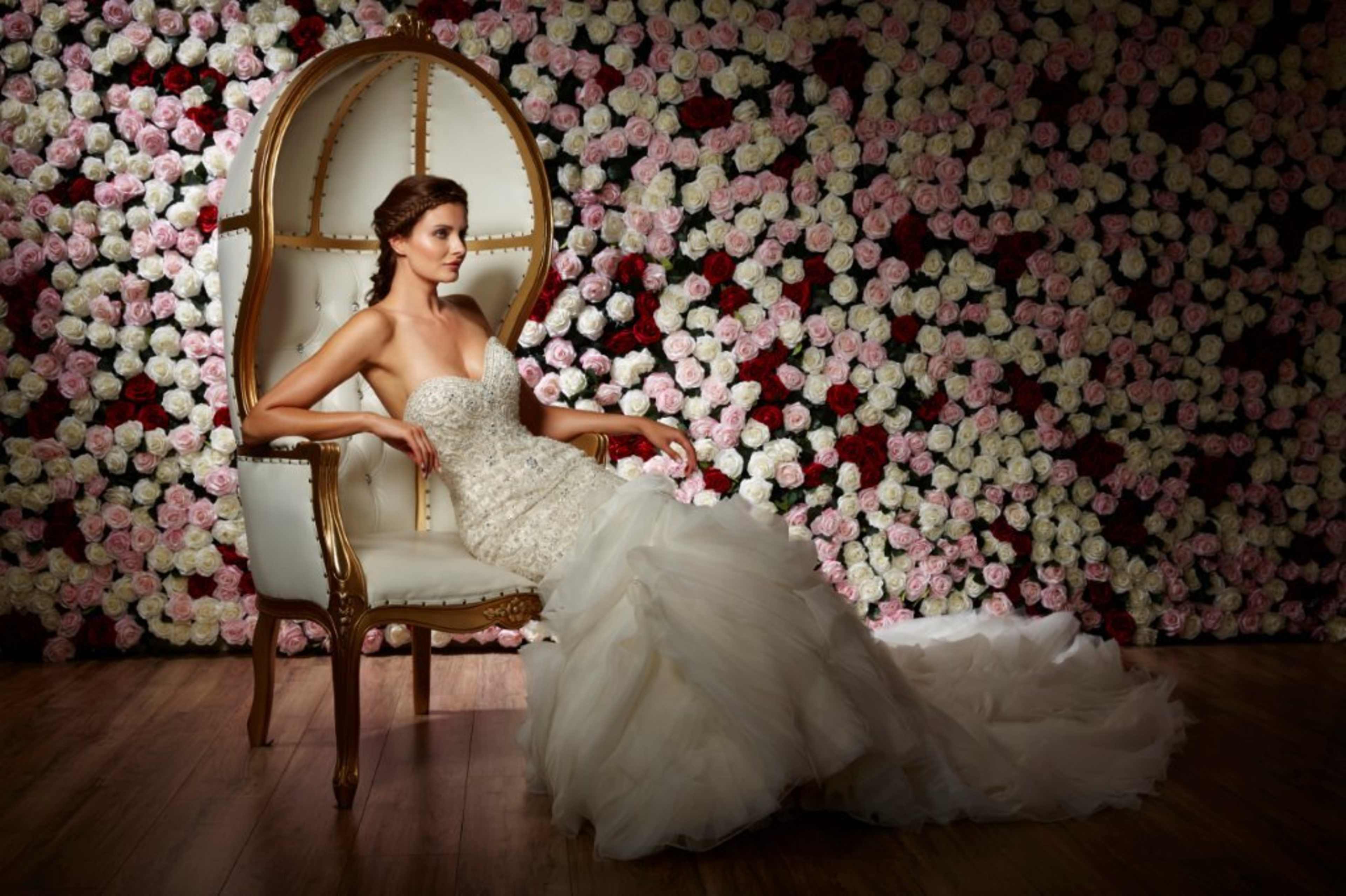 A woman in a wedding dress sits elegantly on a large chair in front of a wall covered with rose flowers.