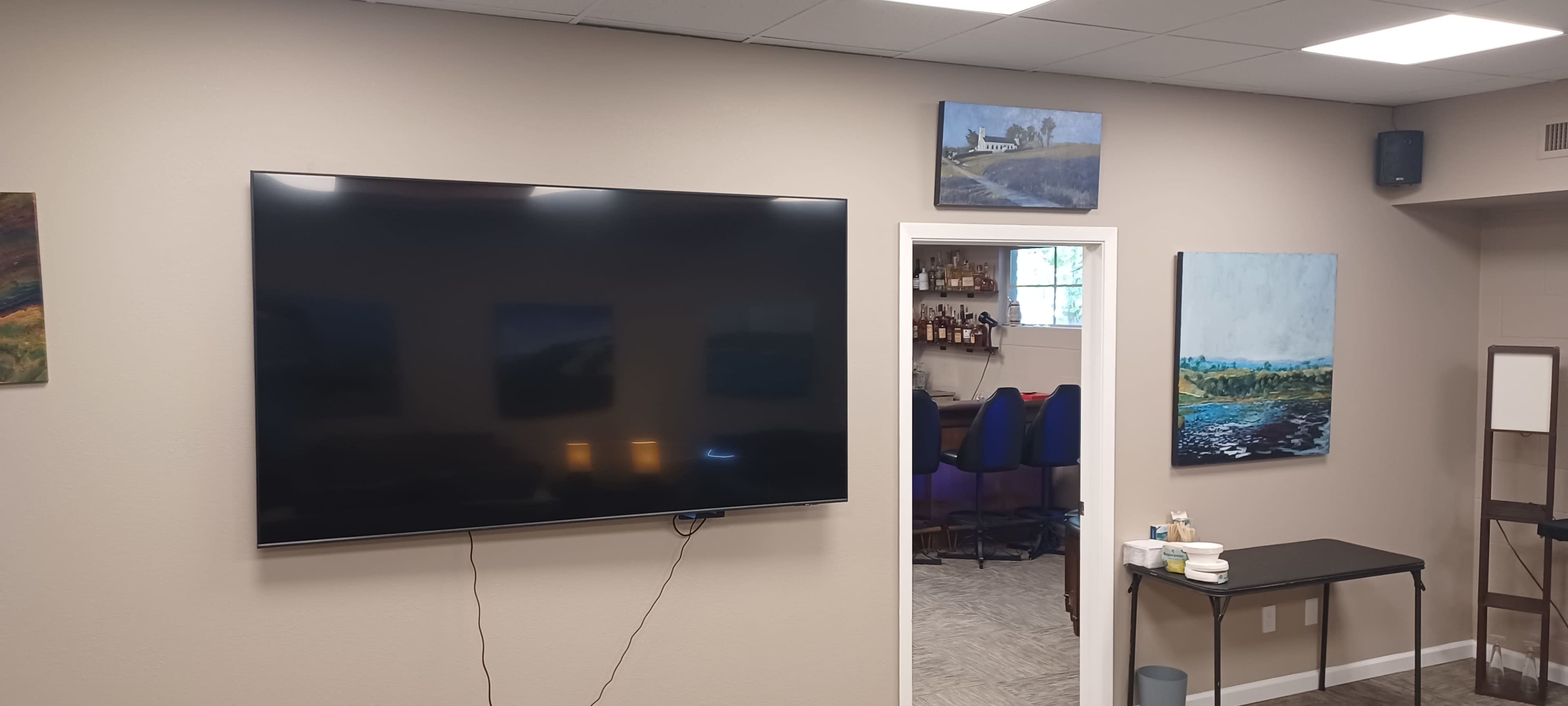 The image shows a room with a large flat-screen TV mounted on a beige wall, surrounded by several paintings, and an adjacent area that includes a bar setup.