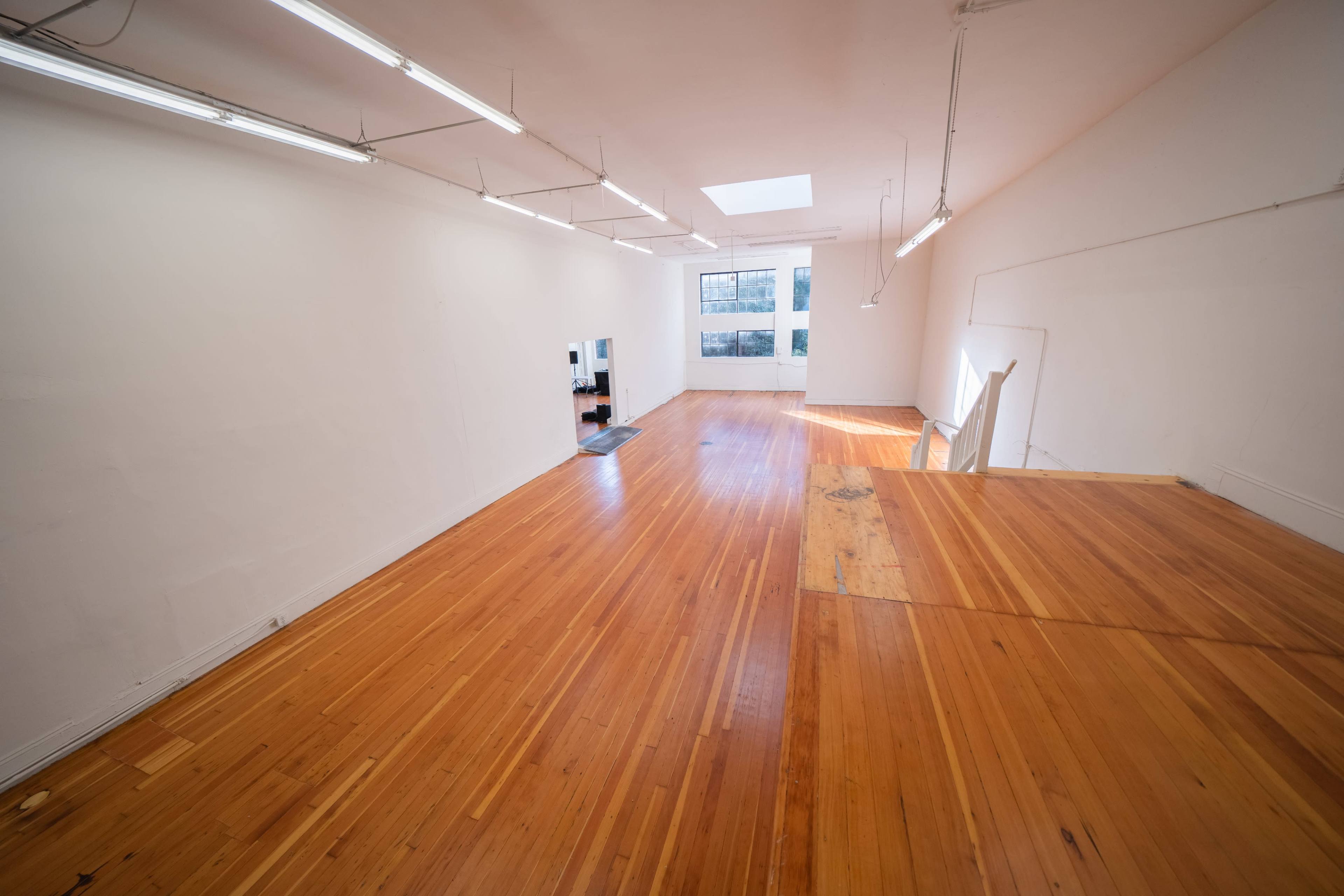 An empty room with wooden flooring, bright overhead lighting, and a skylight, featuring a small area with a black and white appliance setup.