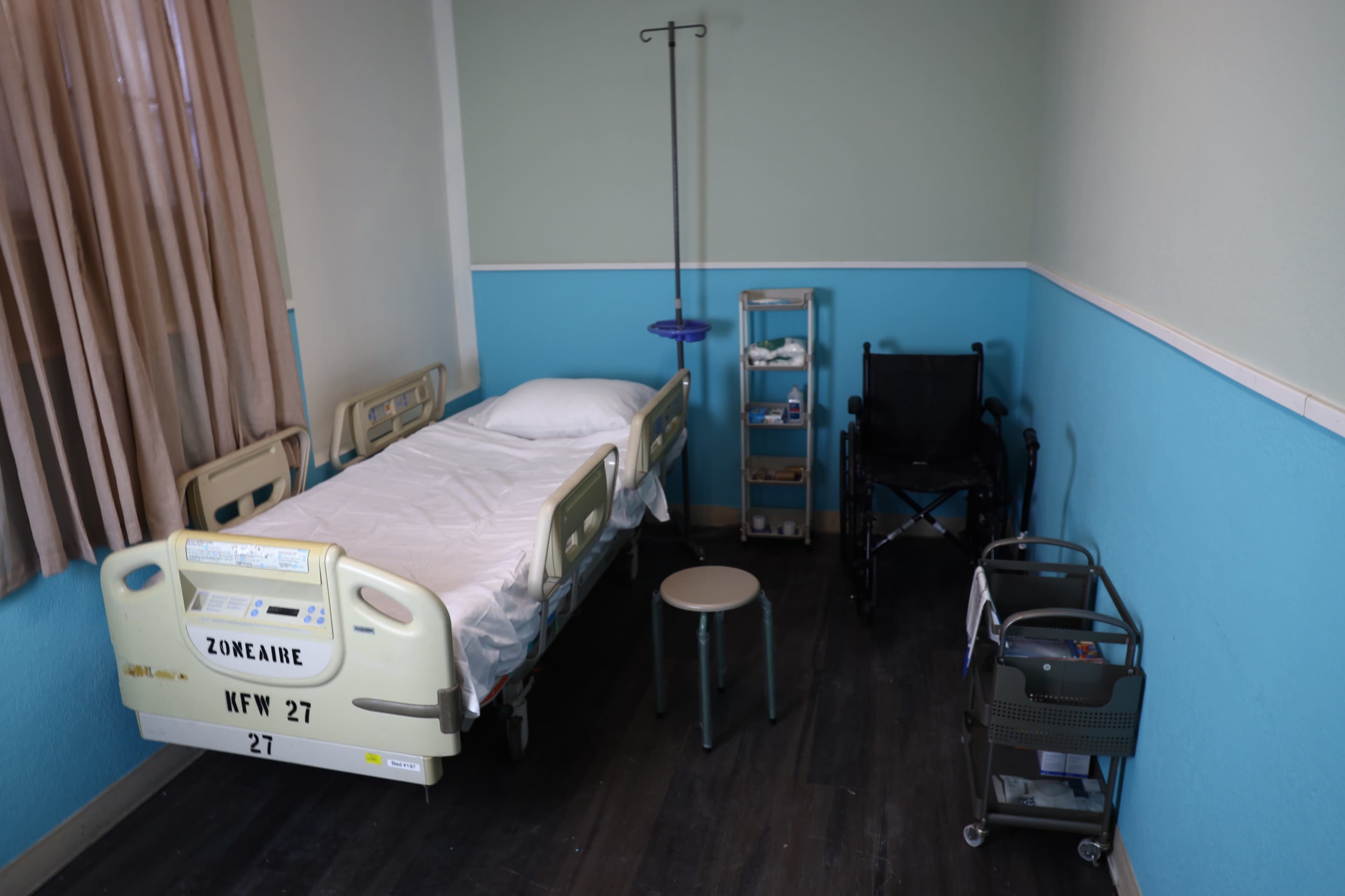The image shows a hospital room with a bed, a wheelchair, a bedside table, and medical supplies organized in a cart.